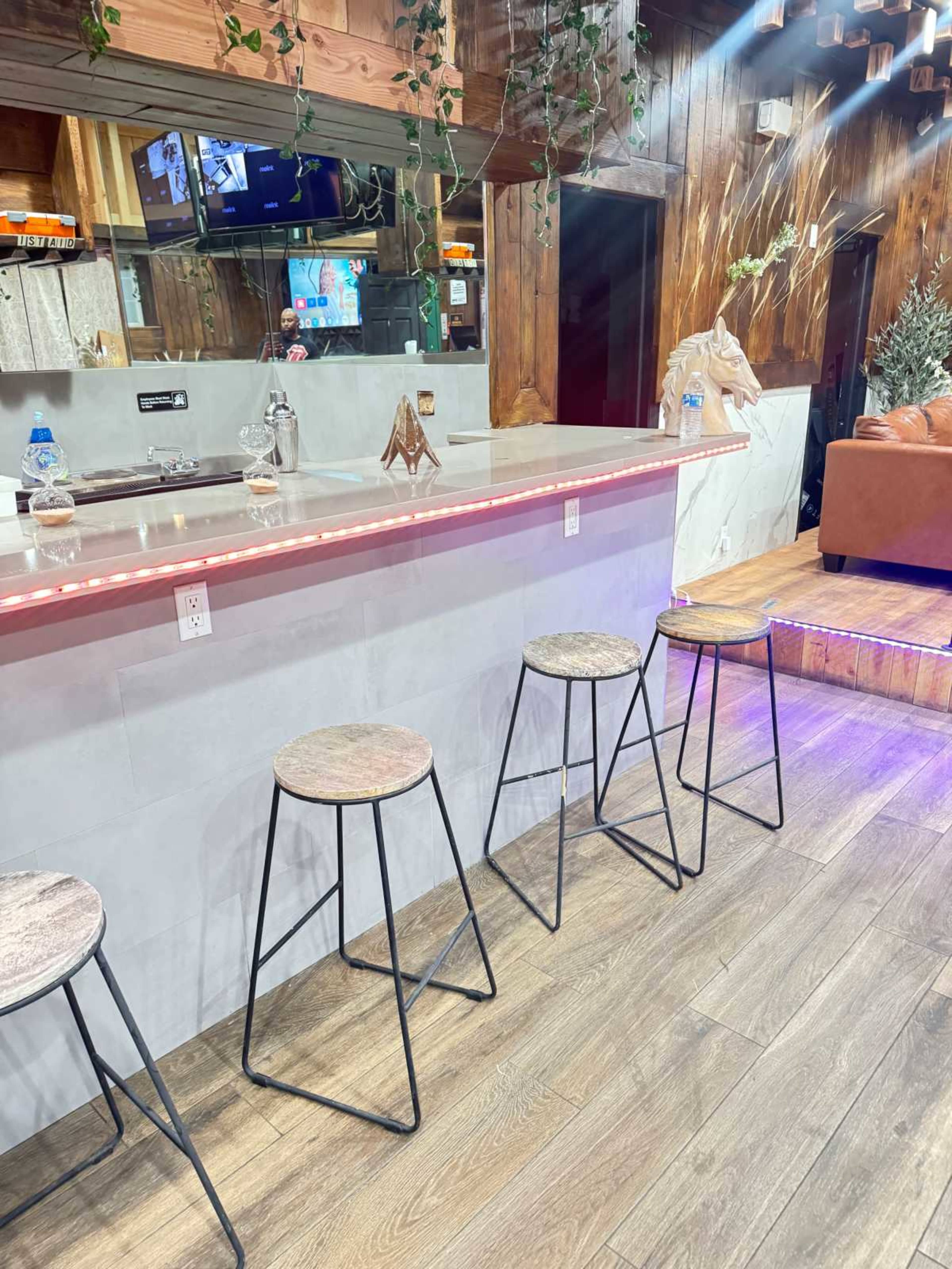 The image shows a modern bar area with four metal and wooden stools positioned in front of a counter, surrounded by wooden paneling and greenery.