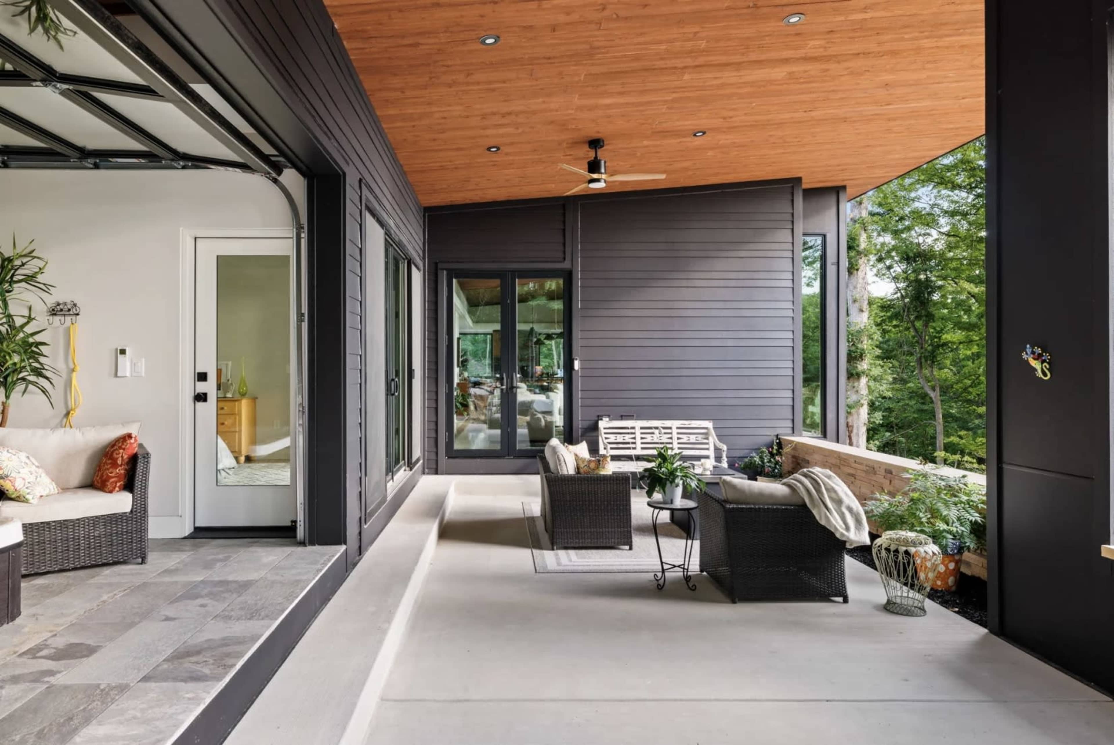 The image shows a modern outdoor living space with a covered patio, featuring comfortable seating, potted plants, and an adjoining garage entrance.