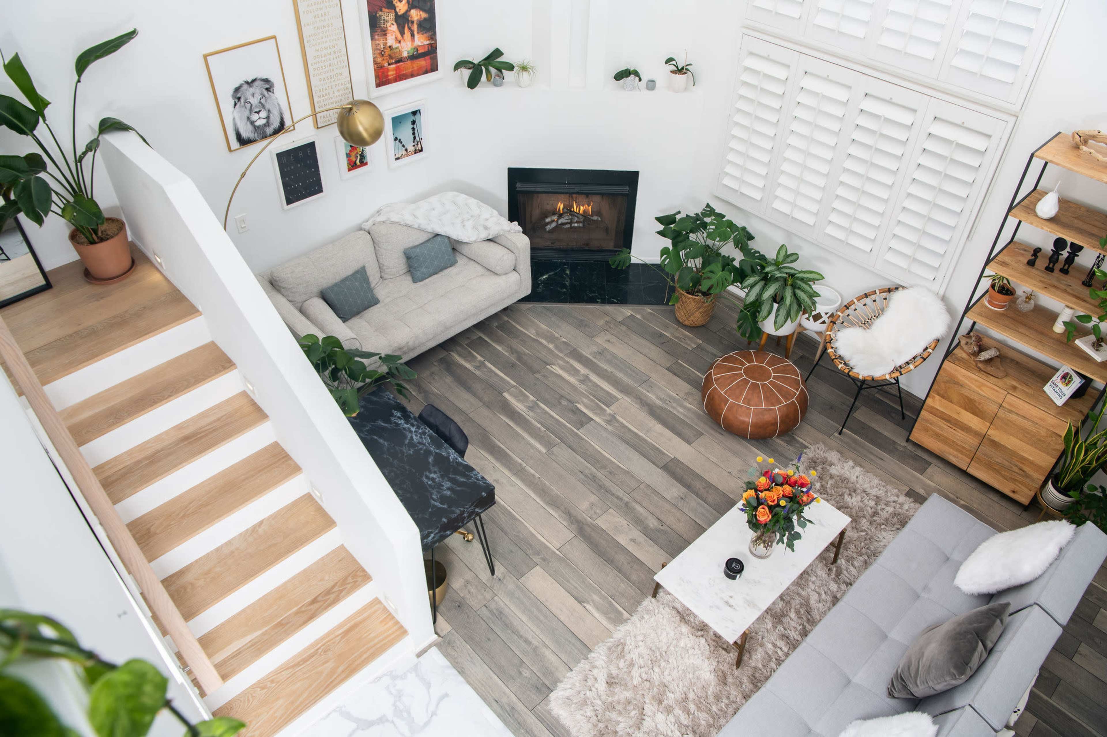 The cozy living room features a gray sofa, a coffee table on a light rug, a fireplace, and various indoor plants, with wooden stairs leading to the upper level.