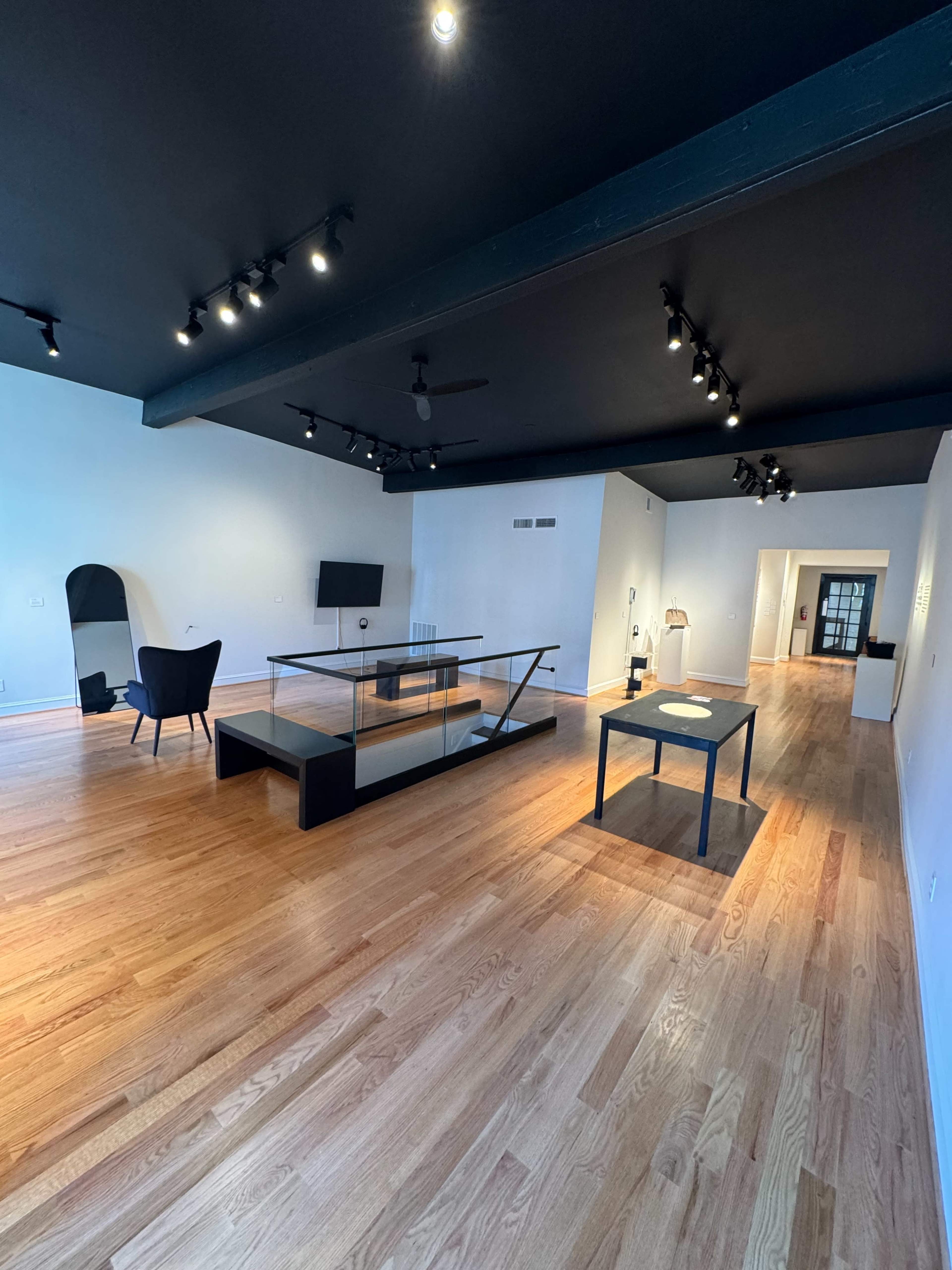 The image shows a spacious room with wooden flooring, featuring a glass-topped table, a black chair, and modern lighting against a dark ceiling.
