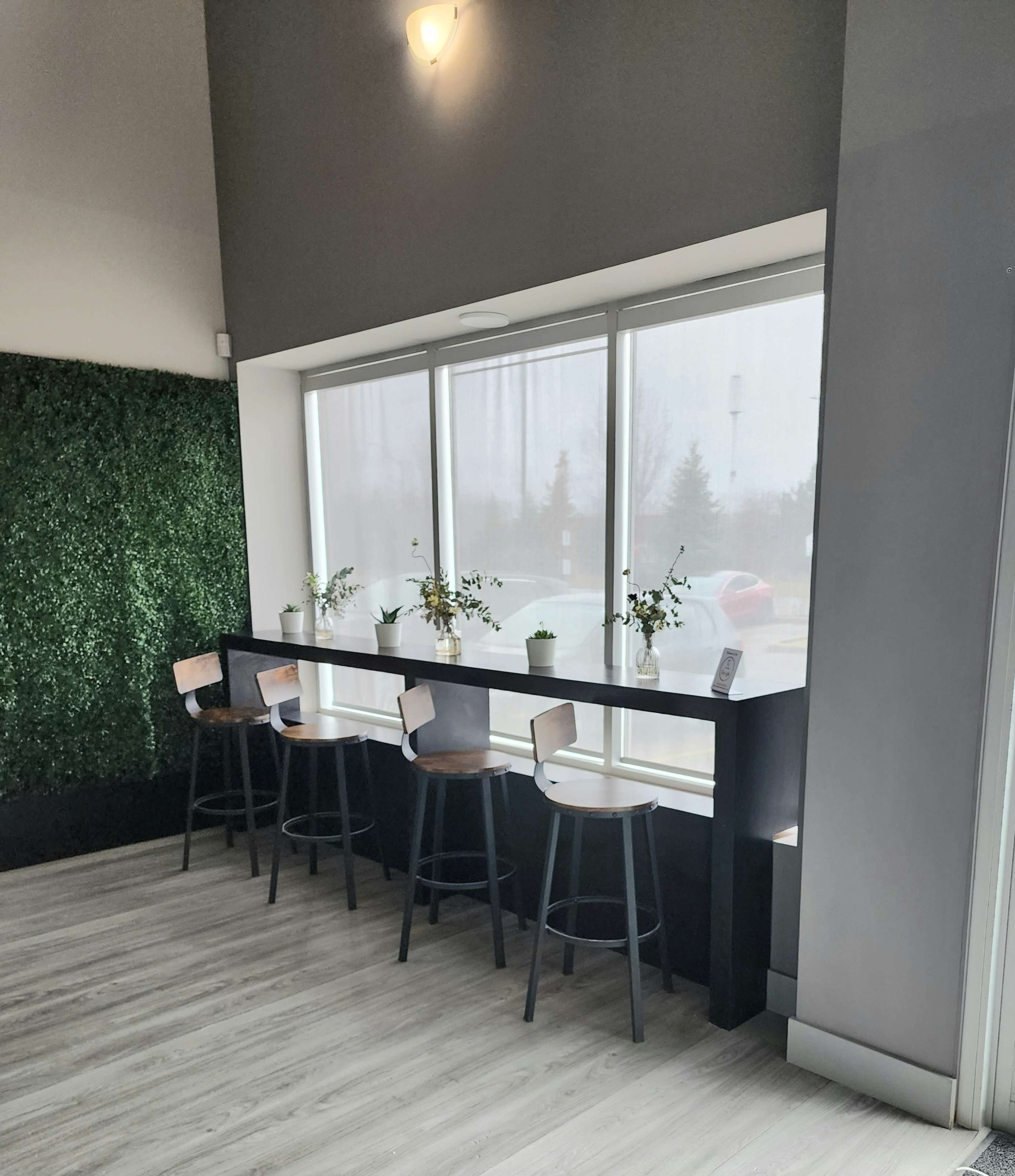 The image shows a modern interior space with a window-side seating area featuring a long table and four stools, surrounded by potted plants and a green wall.