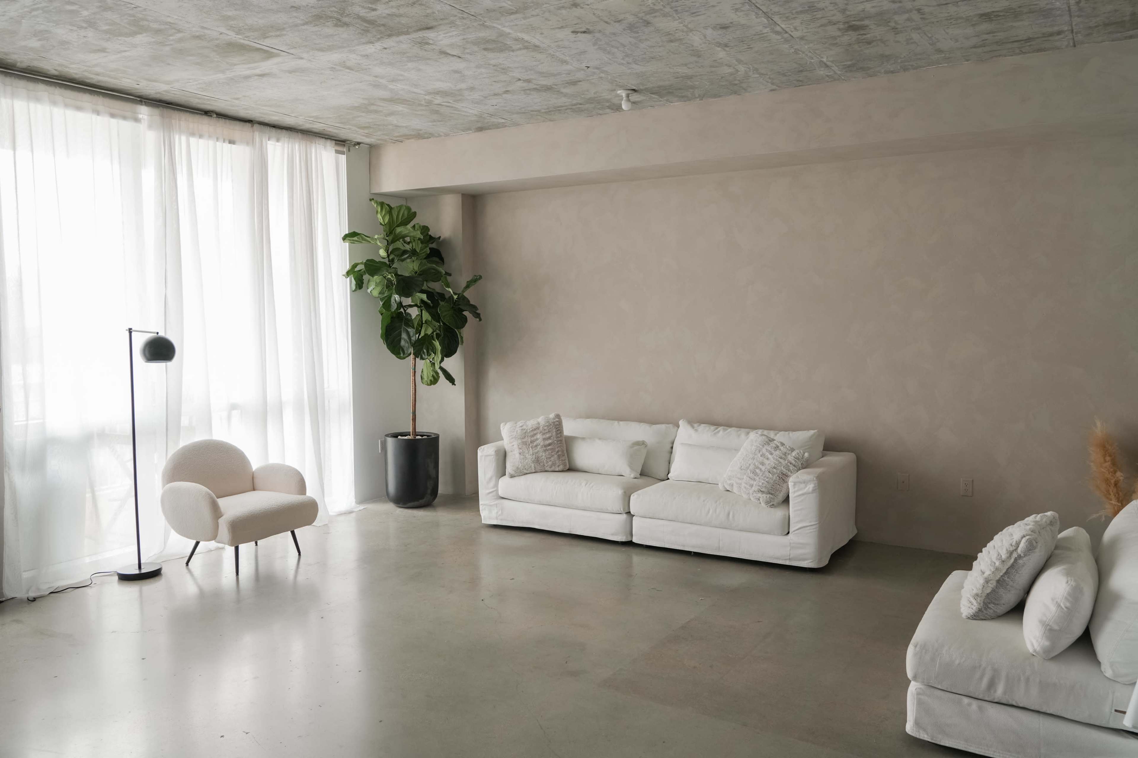A spacious living room features white sofas, a small chair, a floor lamp, and a potted plant, with large windows allowing natural light to enter.