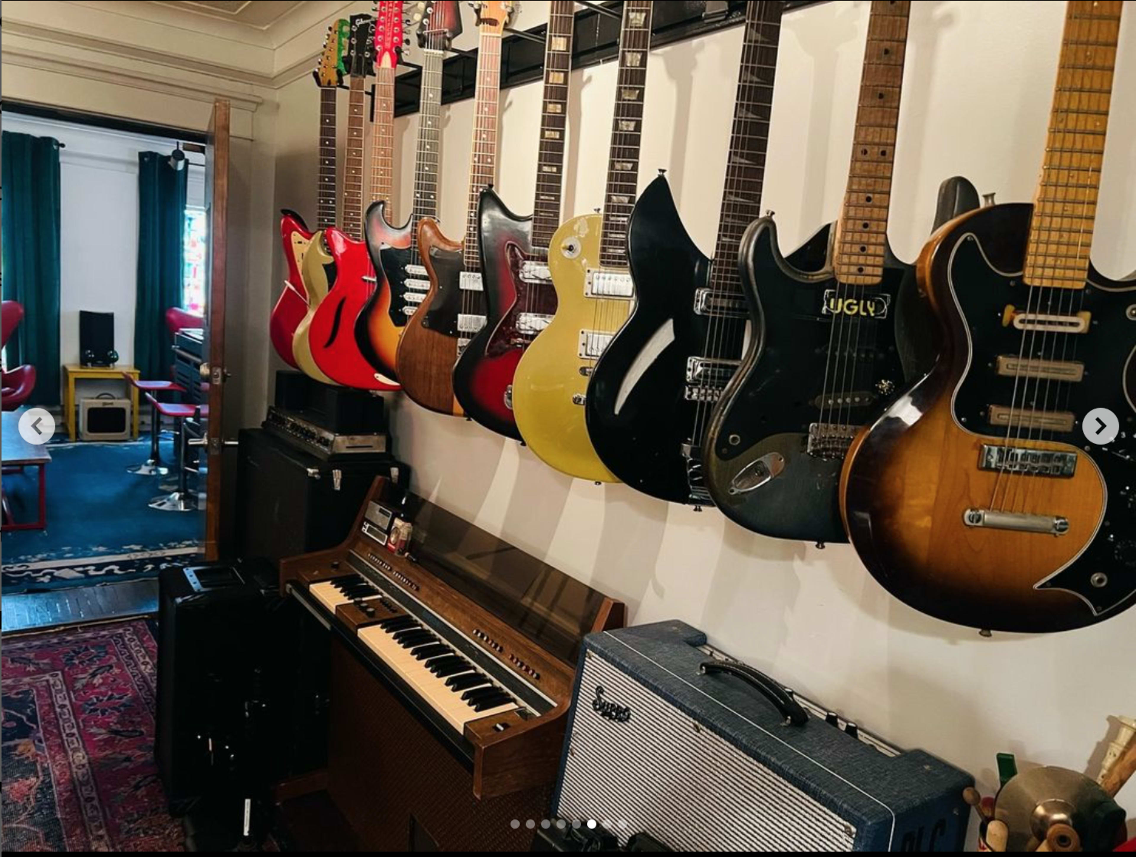 A variety of guitars are displayed on a wall next to a vintage keyboard and amplifiers in a room with a red chair and green curtains visible in the background.