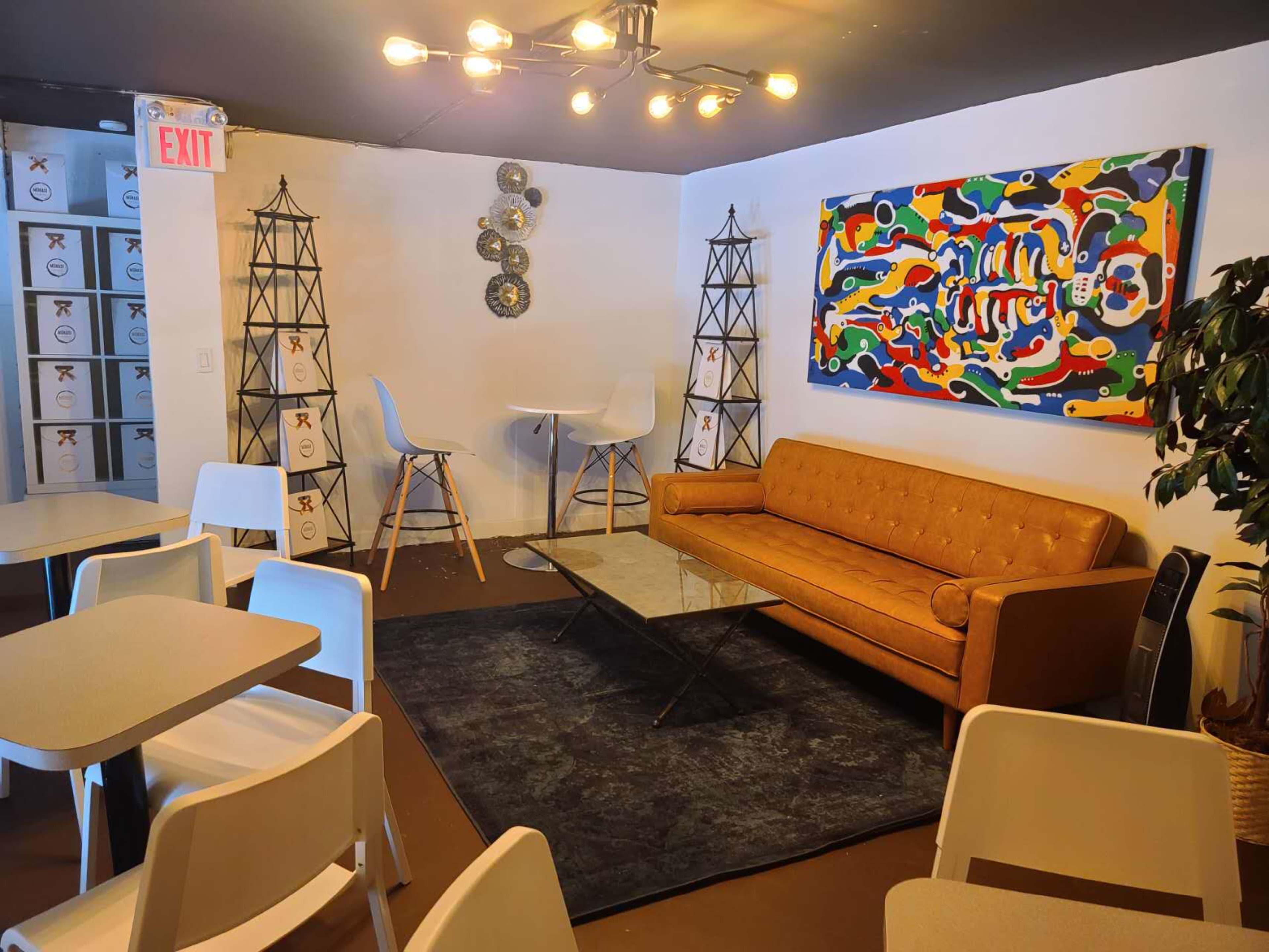 A cozy seating area with a brown sofa, a modern table with white chairs, and vibrant artwork on the wall, surrounded by light fixtures and decorative elements.