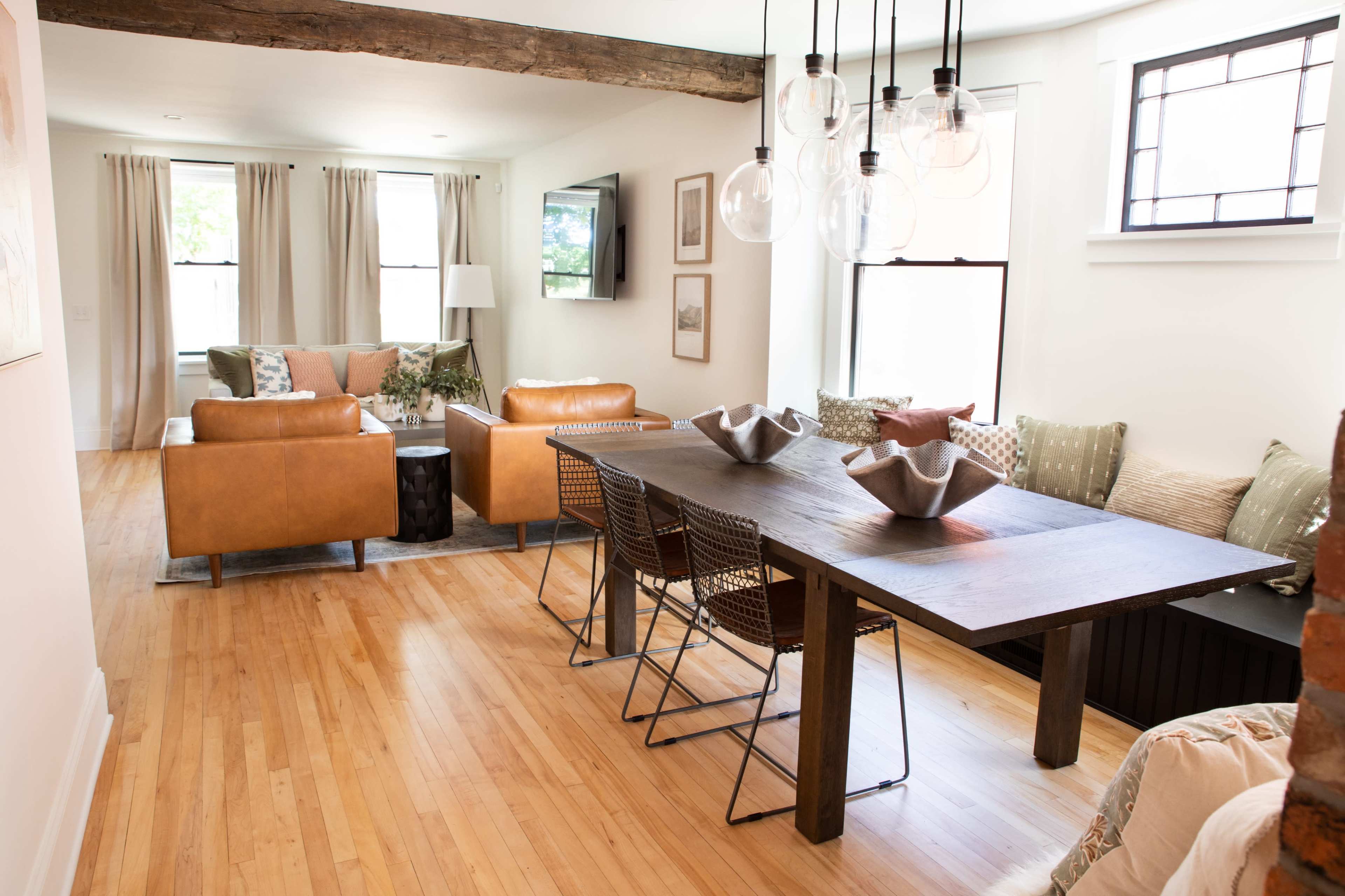 A modern dining area with a wooden table and wire chairs, adjacent to a living space featuring leather sofas and large windows.