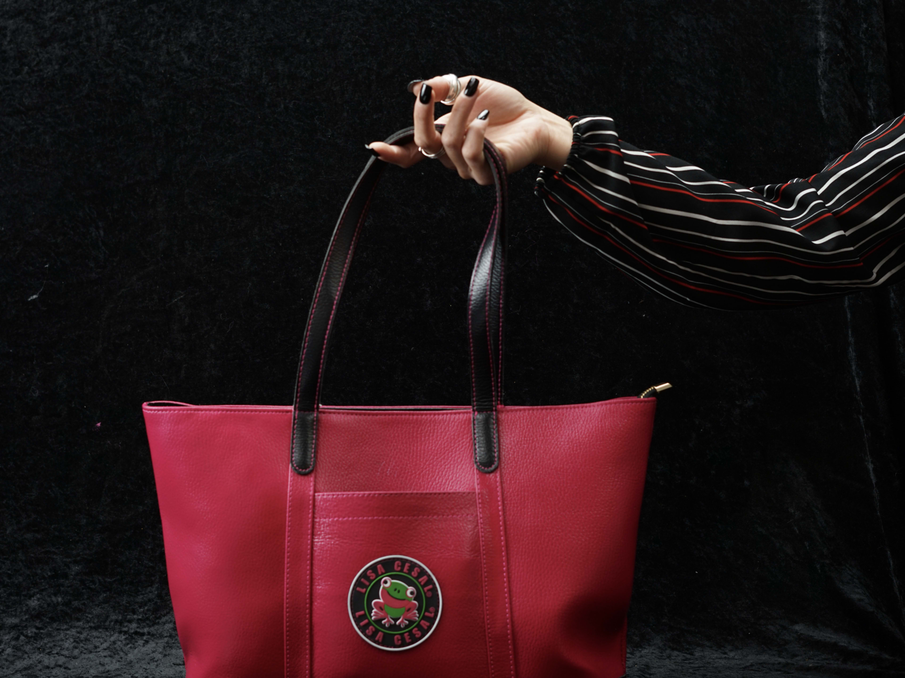 A hand is holding a vibrant pink tote bag featuring a circular logo, against a dark background.