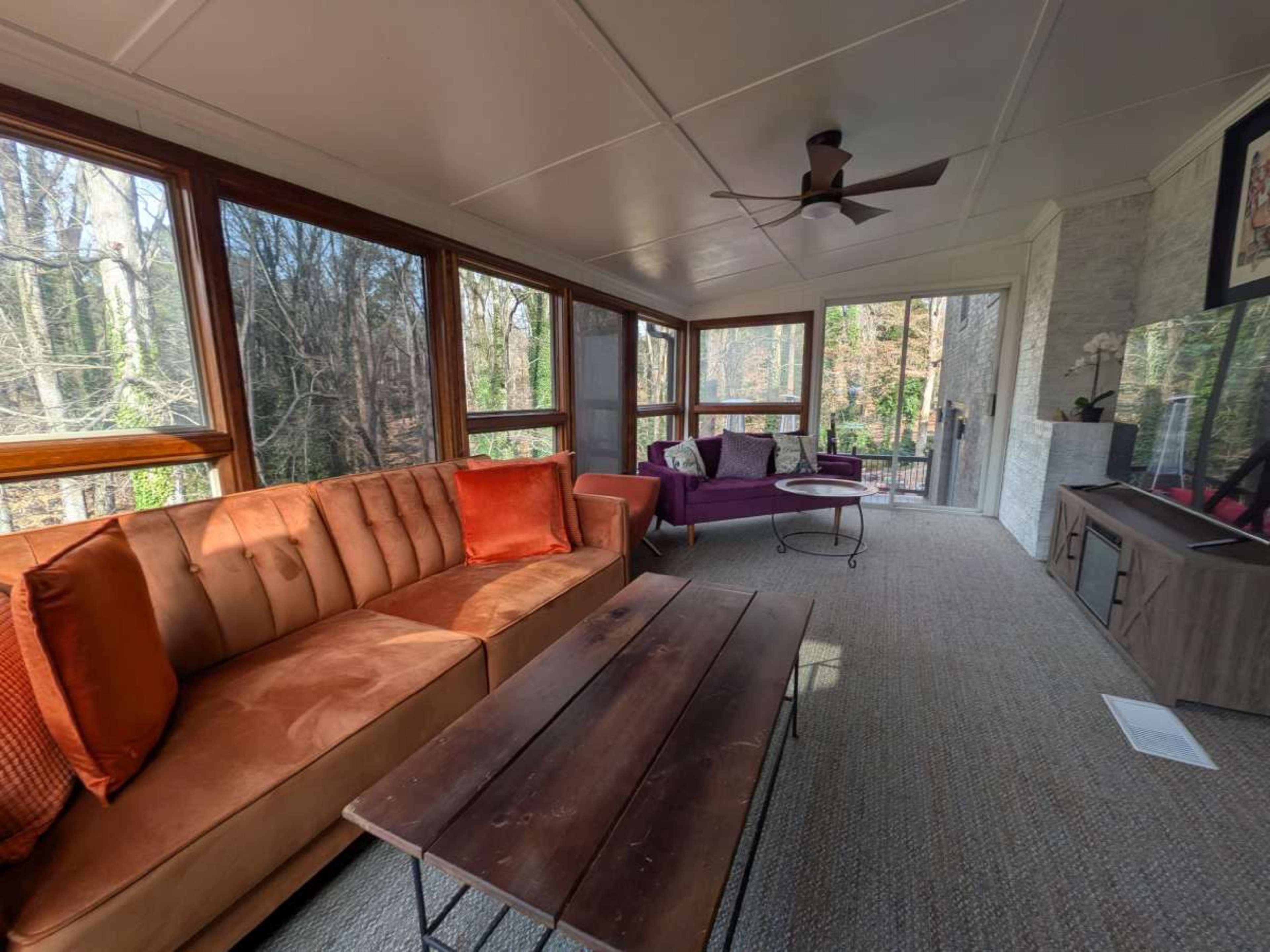 The image shows a sunlit screened-in porch featuring a brown sofa, a purple couch, a wooden coffee table, and a view of trees outside.