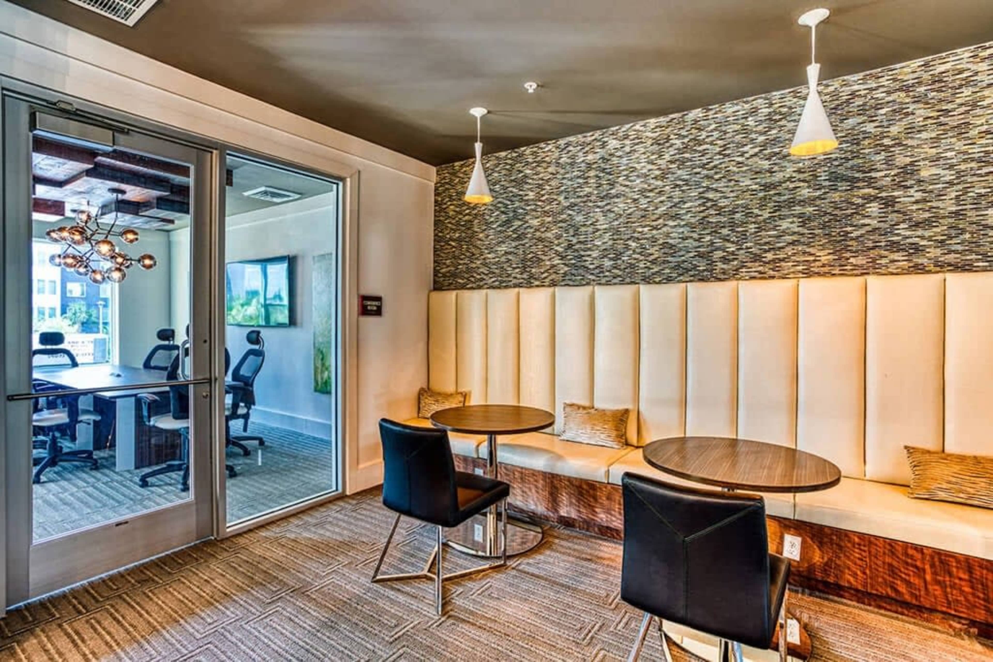 The image shows a modern seating area with two small round tables and black chairs, adjacent to a glass door leading to a conference room with chairs around a large table.