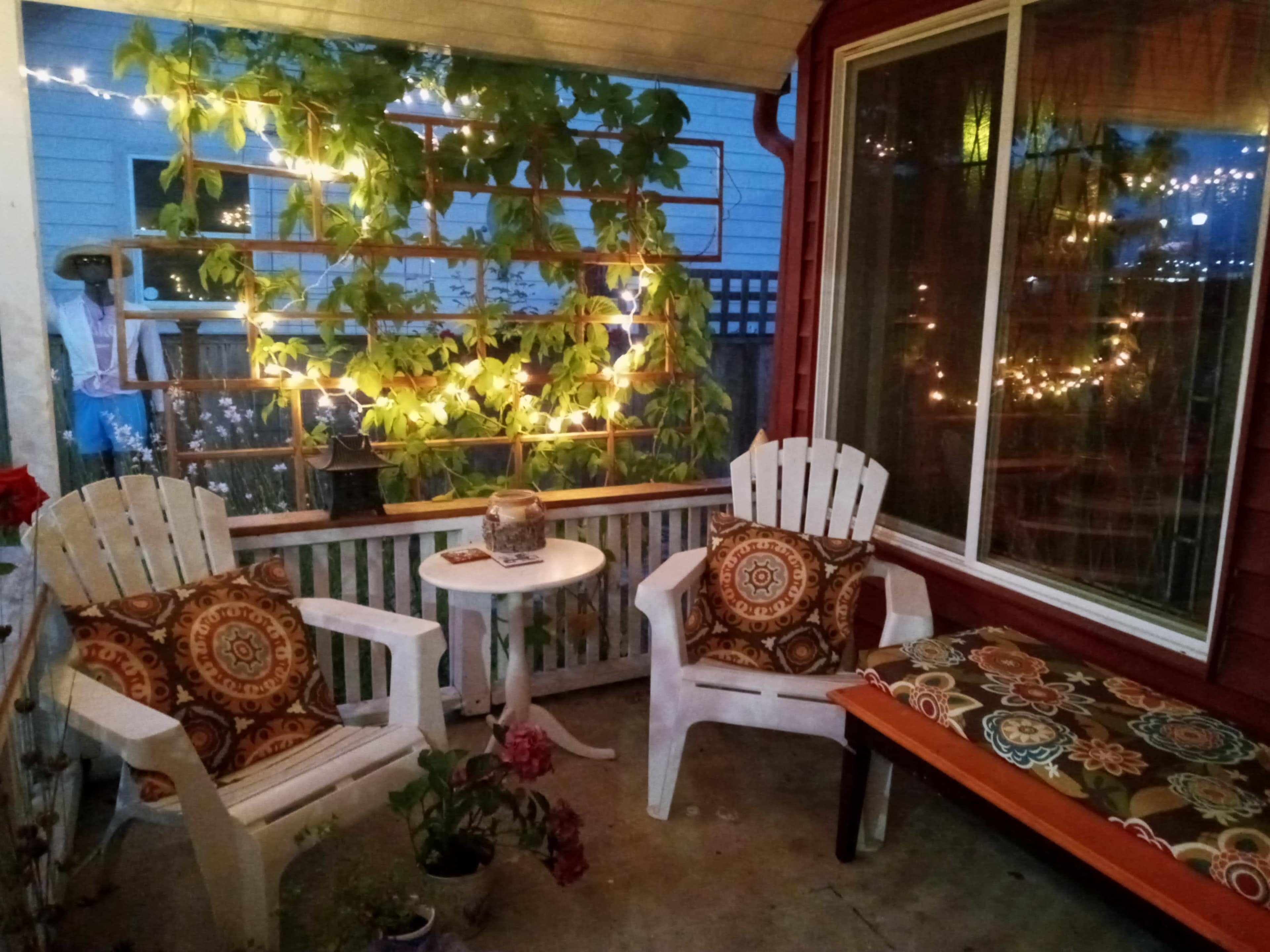 A cozy porch features two white adirondack chairs with patterned cushions, a small table, and greenery illuminated by string lights.