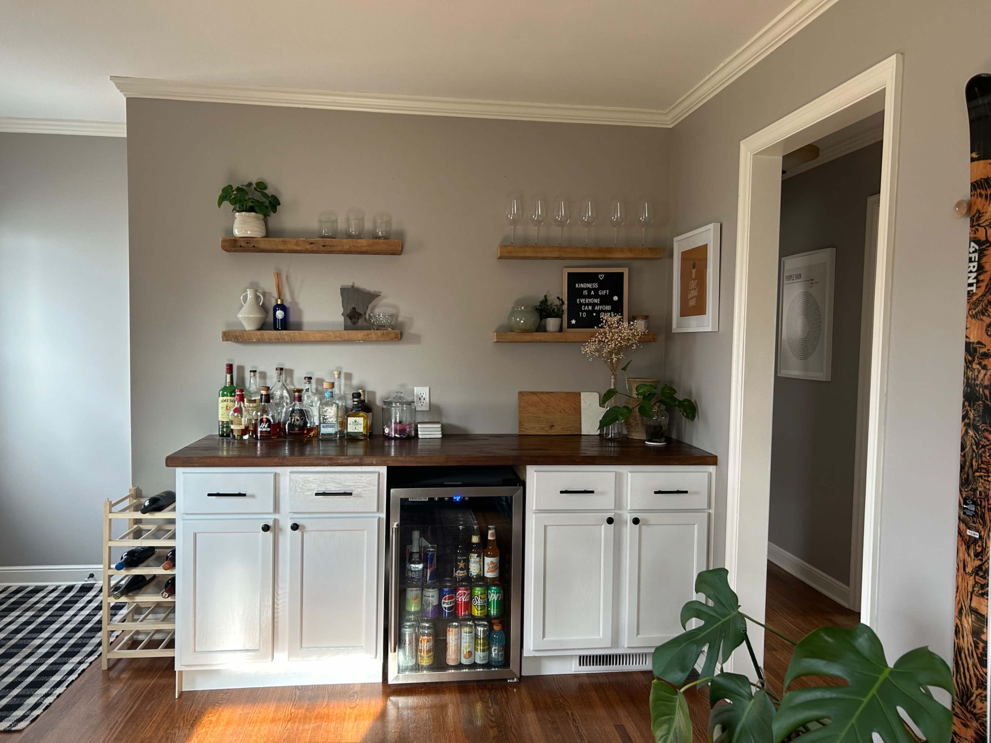 The image shows a home bar area with shelves holding glasses and various bottles of alcohol, accompanied by a mini fridge and a wine rack.