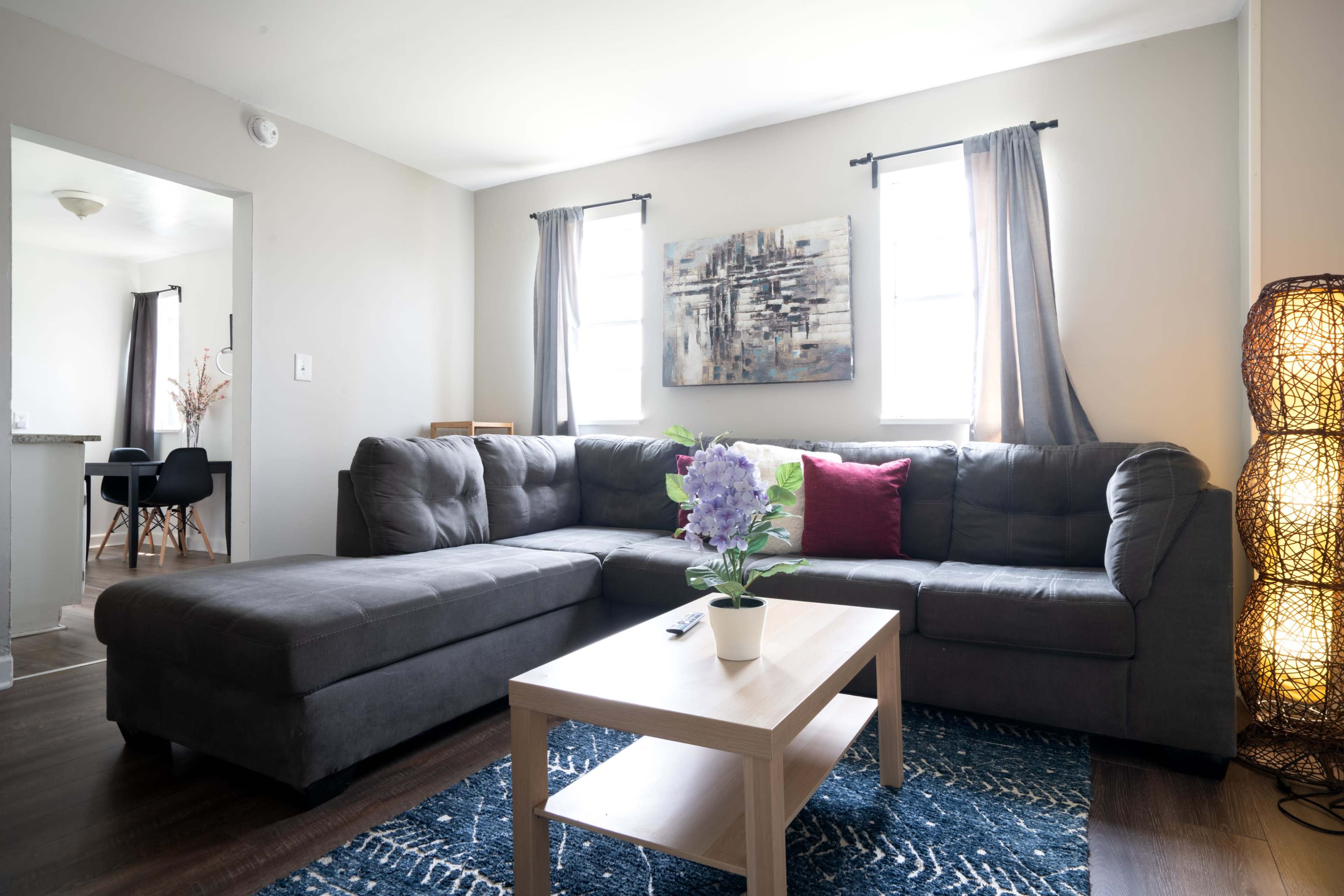 A modern living room features a grey sectional sofa, a wooden coffee table with a potted plant, and a decorative area rug, illuminated by natural light from two windows.