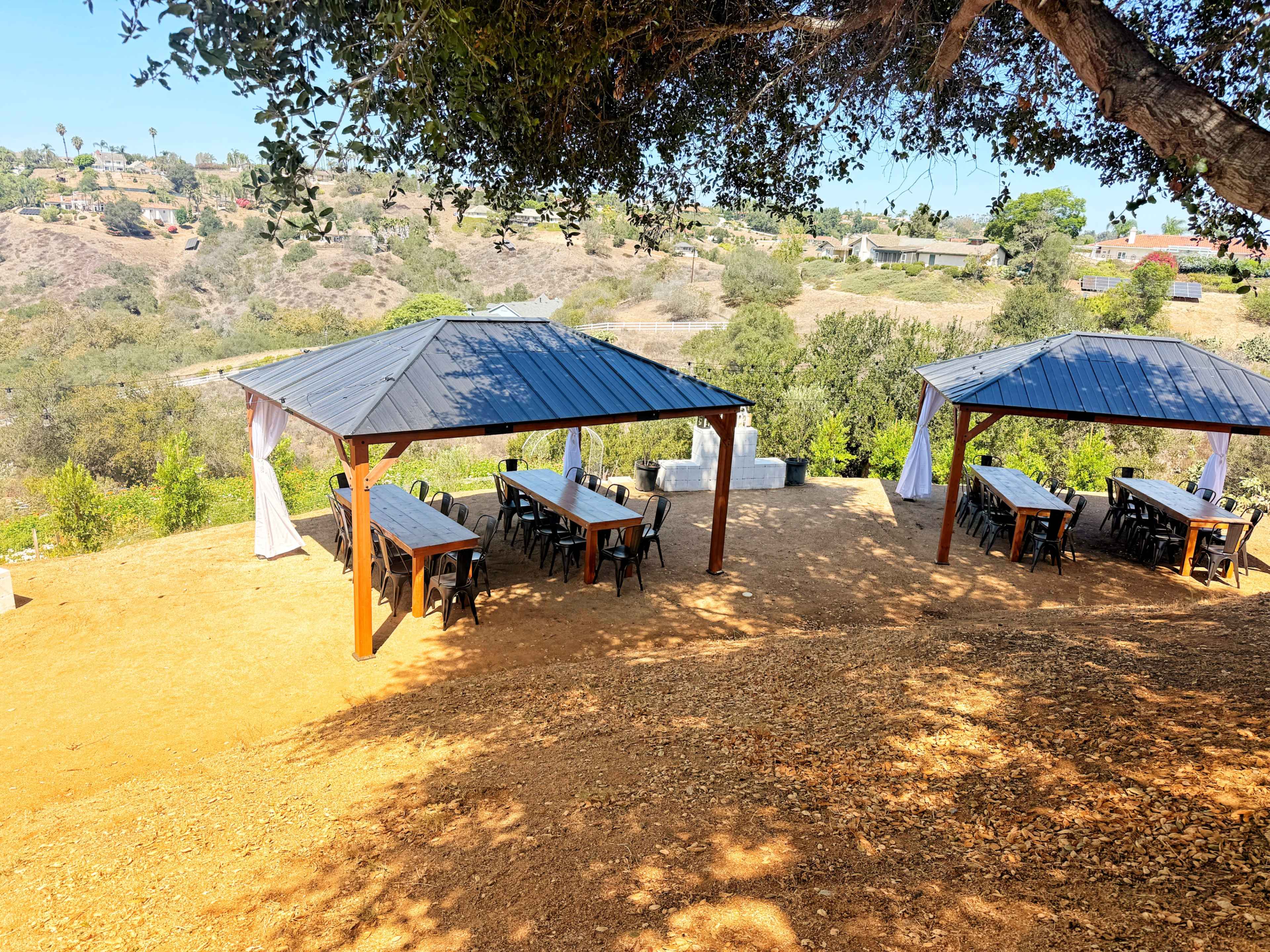 The image shows a landscaped outdoor area with two covered seating pavilions and multiple tables set under them, surrounded by greenery and hills in the background.
