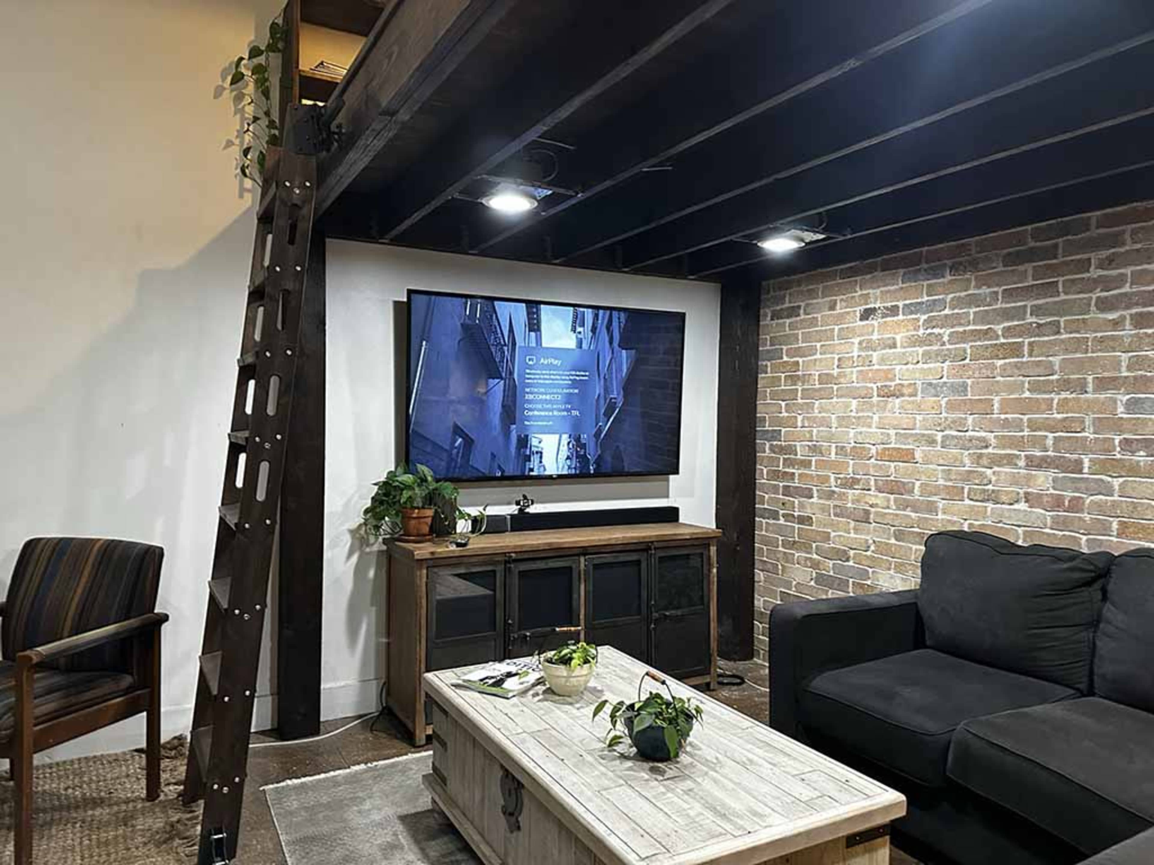 A basement living area featuring a dark sofa, a wooden coffee table with plants, a television mounted on the wall, and exposed brick alongside wooden beams.