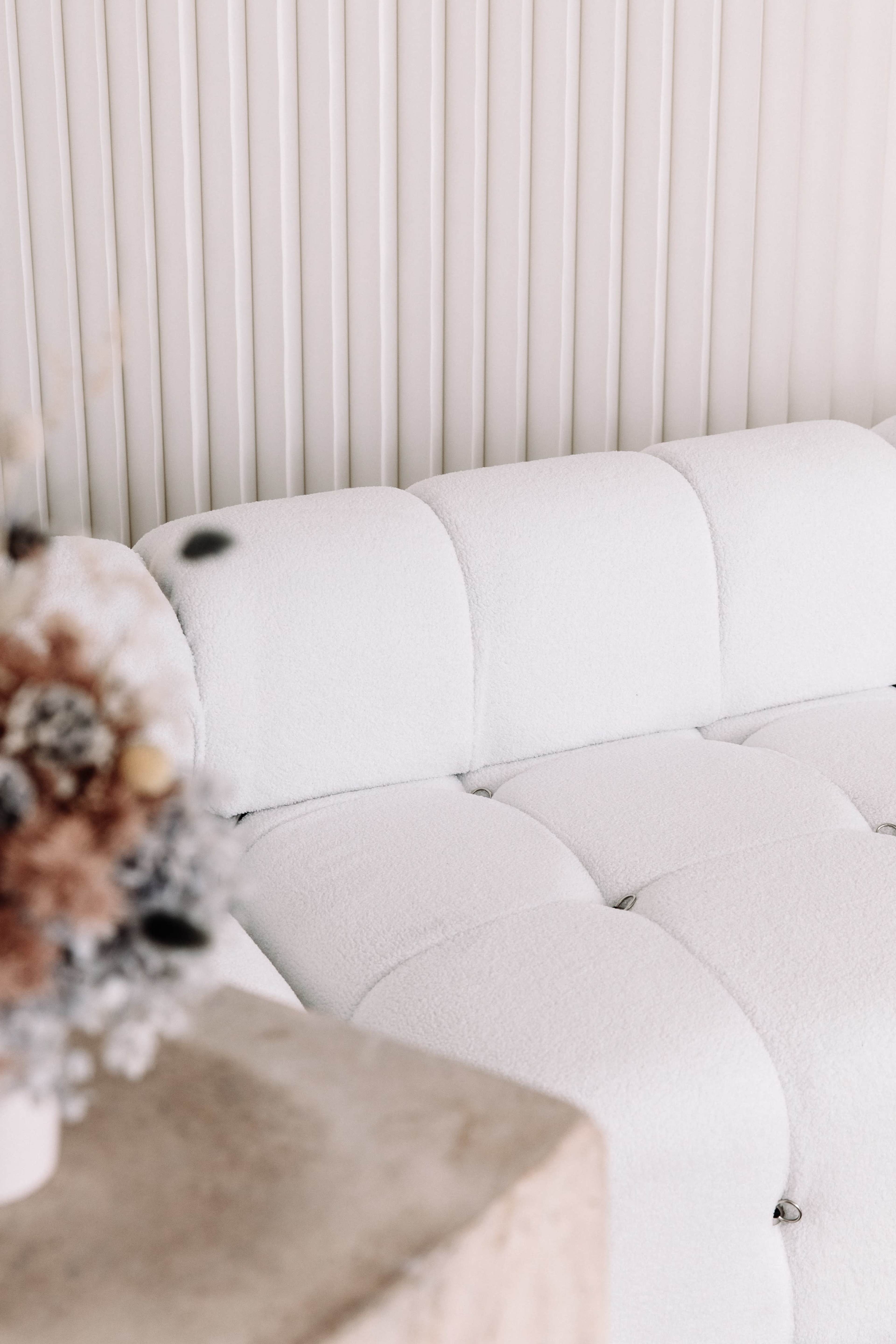 A white, tufted sofa is positioned next to a textured beige surface and a decorative arrangement of dried flowers.