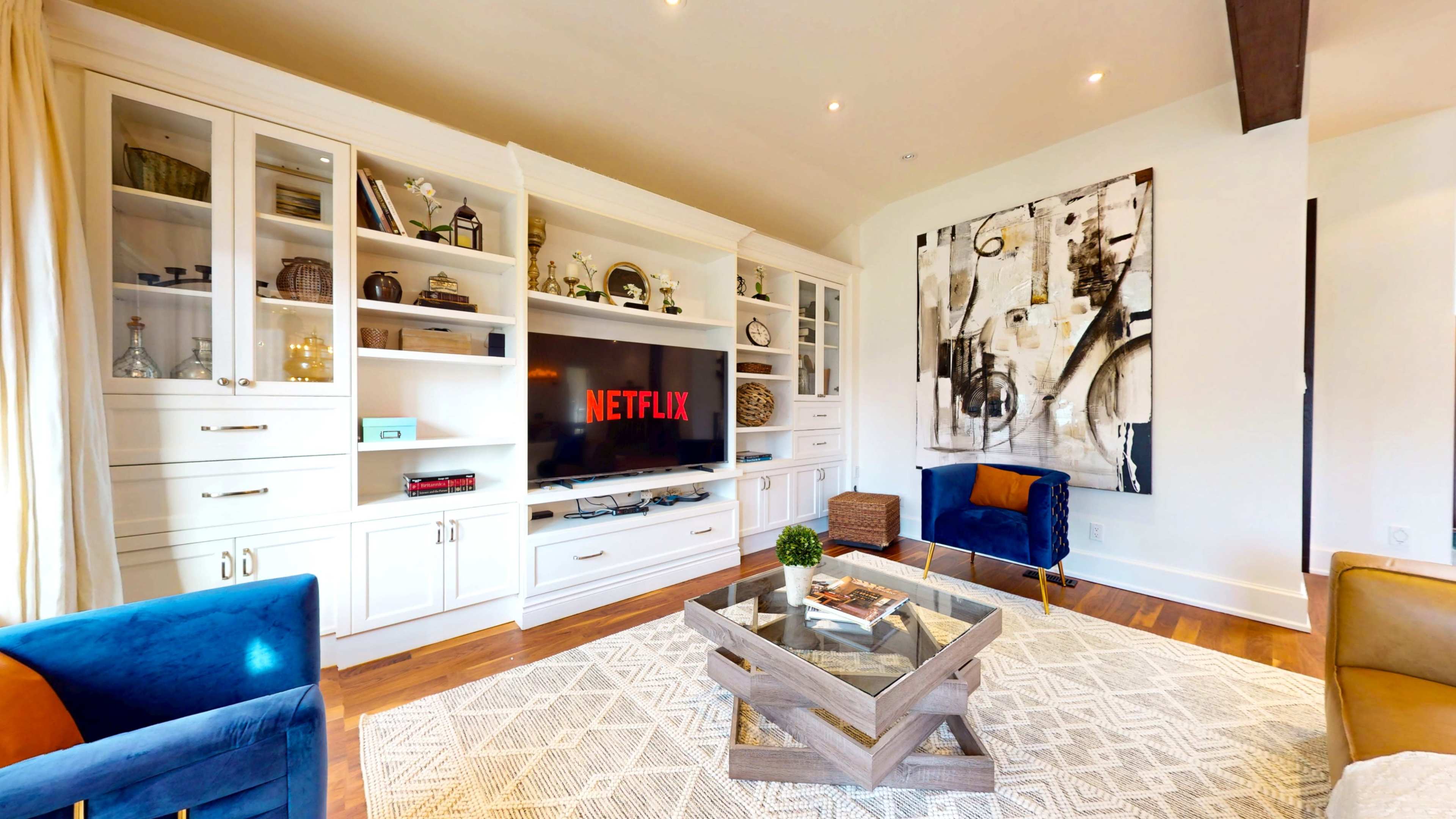 A modern living room features a large TV displaying the Netflix logo, surrounded by built-in shelves, a coffee table, and two accent chairs.