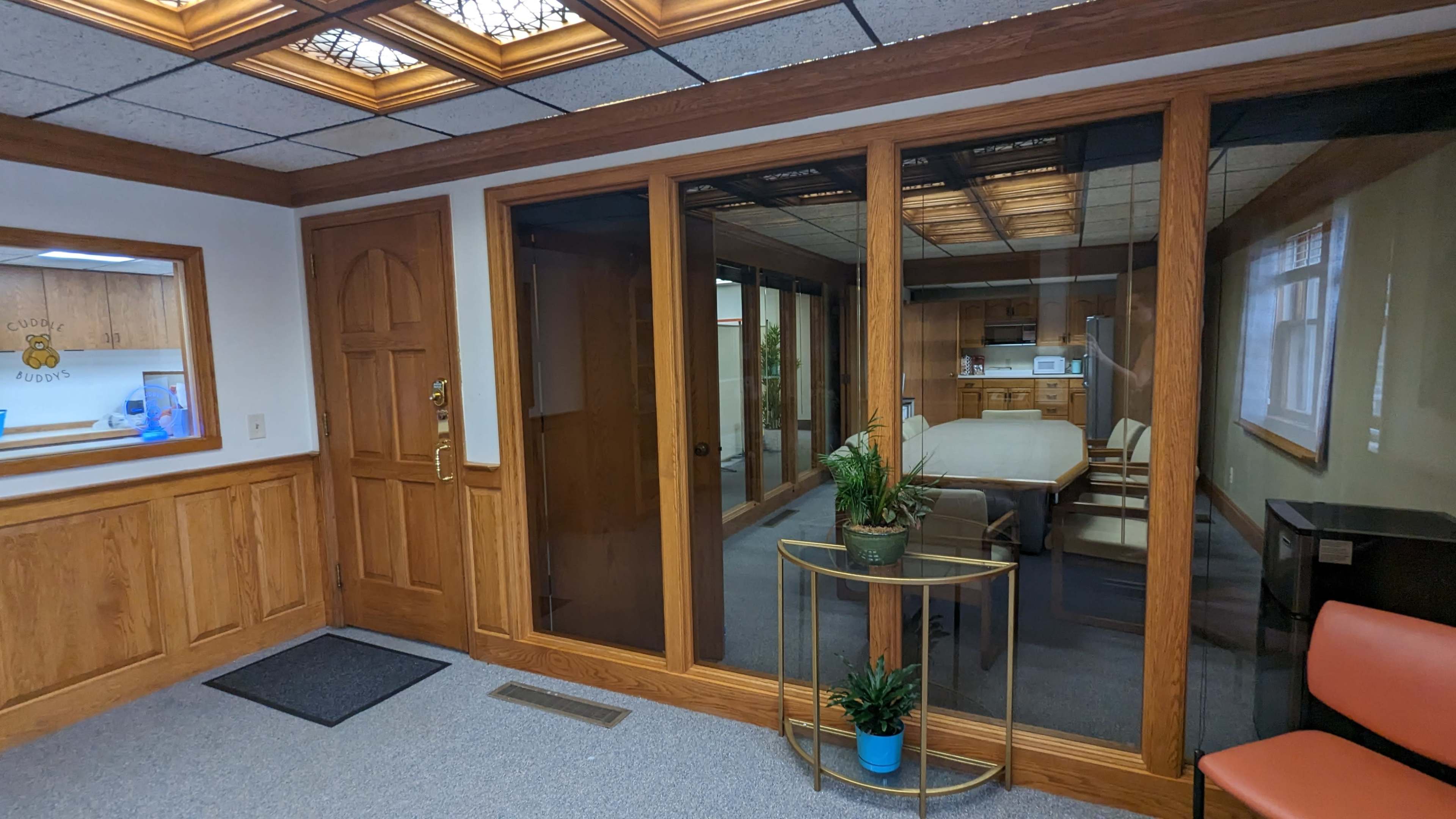 An interior office space with a glass-walled conference room, wooden paneling, and a small table with plants near the entrance.