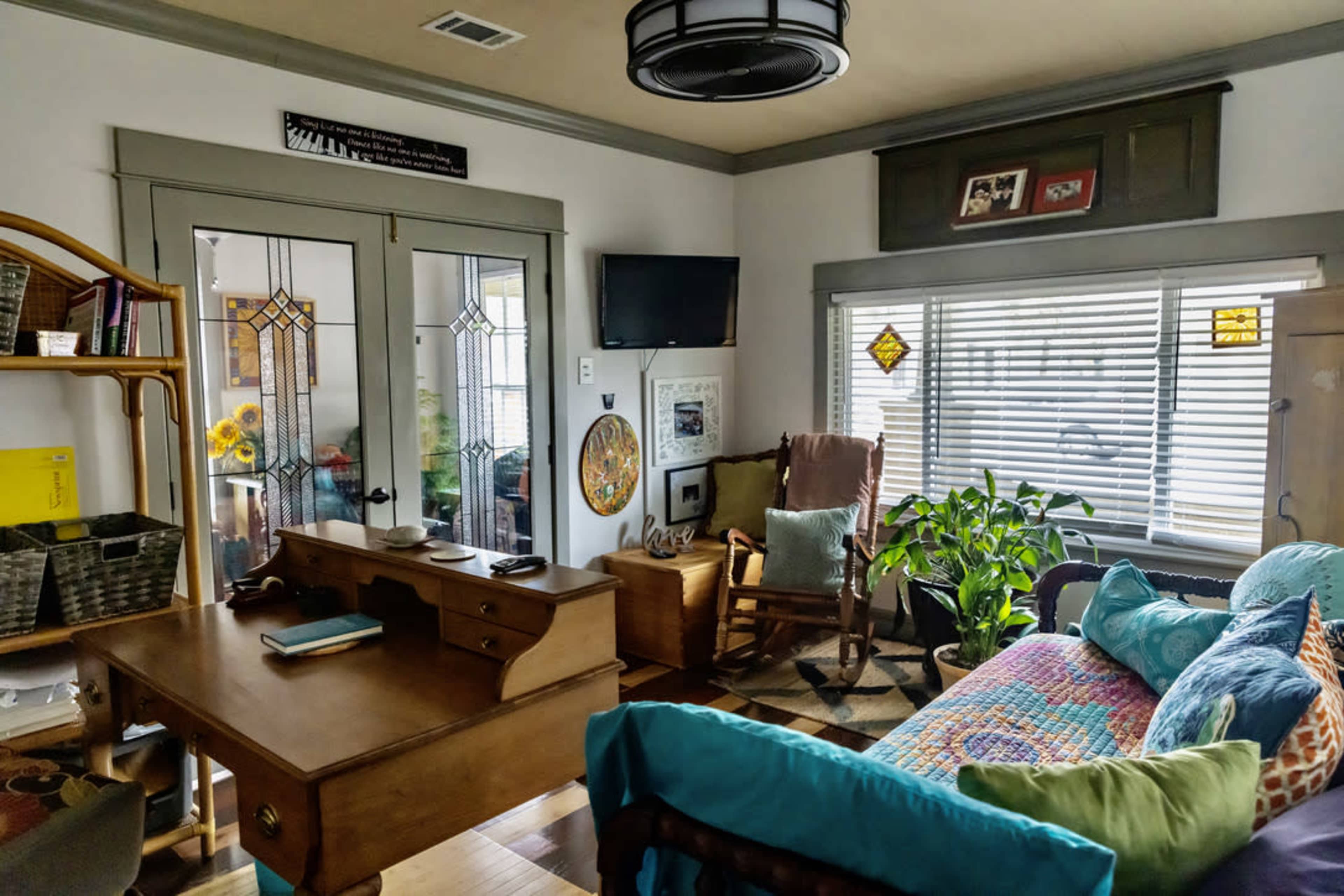 A cozy living room with a wooden desk, a rocking chair, a couch adorned with colorful pillows, and plants near the window.