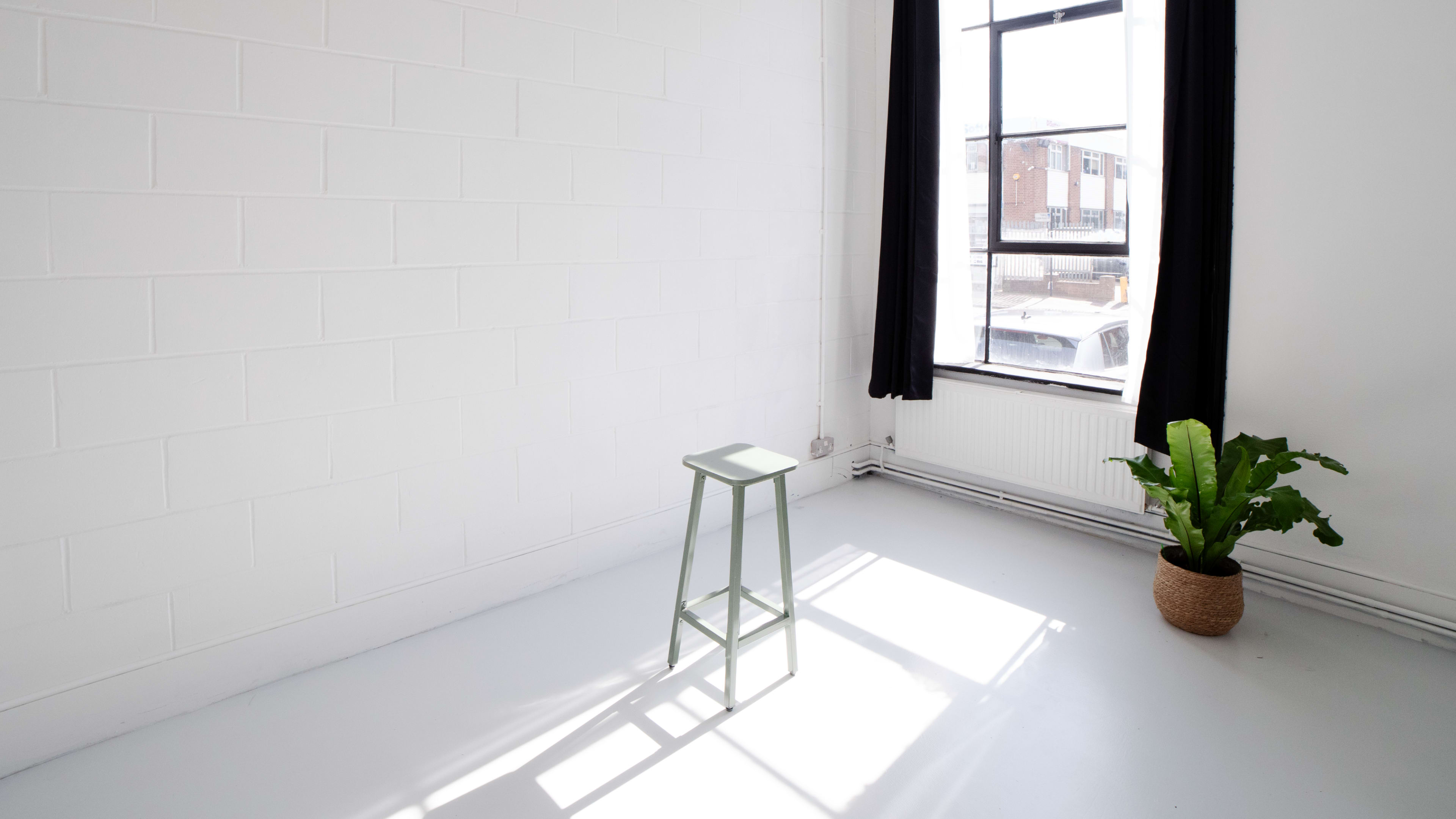 A minimalist room features a green stool next to a large window, with sunlight casting a shadow on the light gray floor.