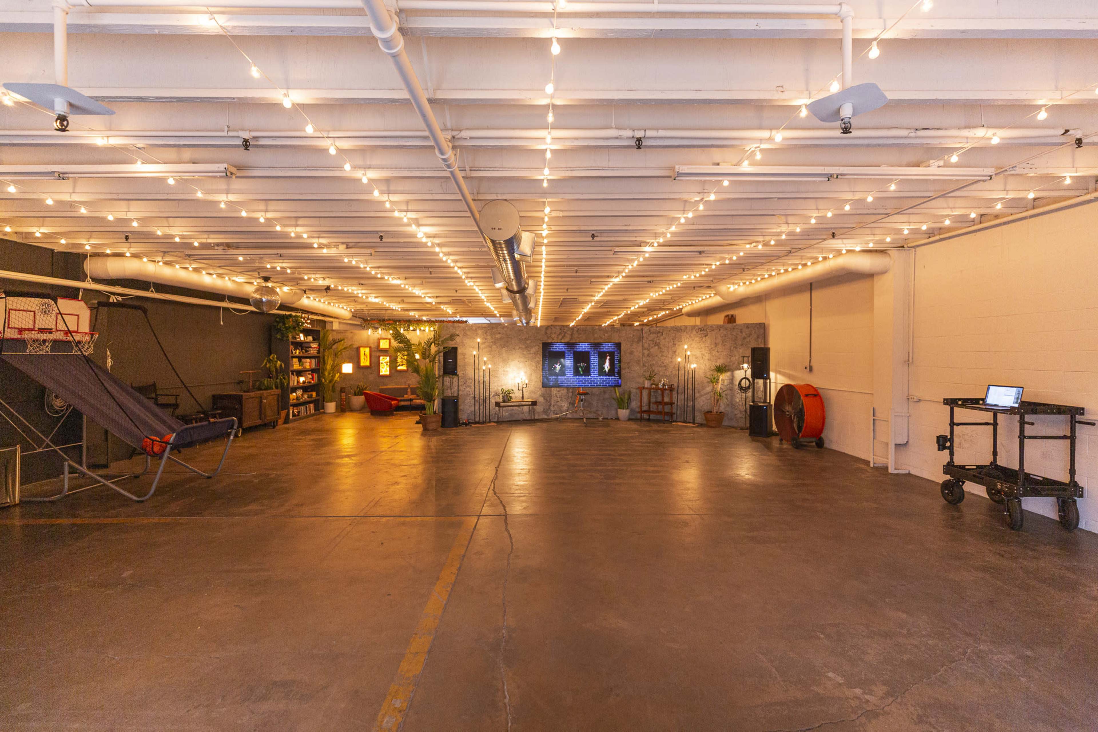 A spacious room with string lights overhead, a hammock, shelving units, and a television setup against a textured wall.