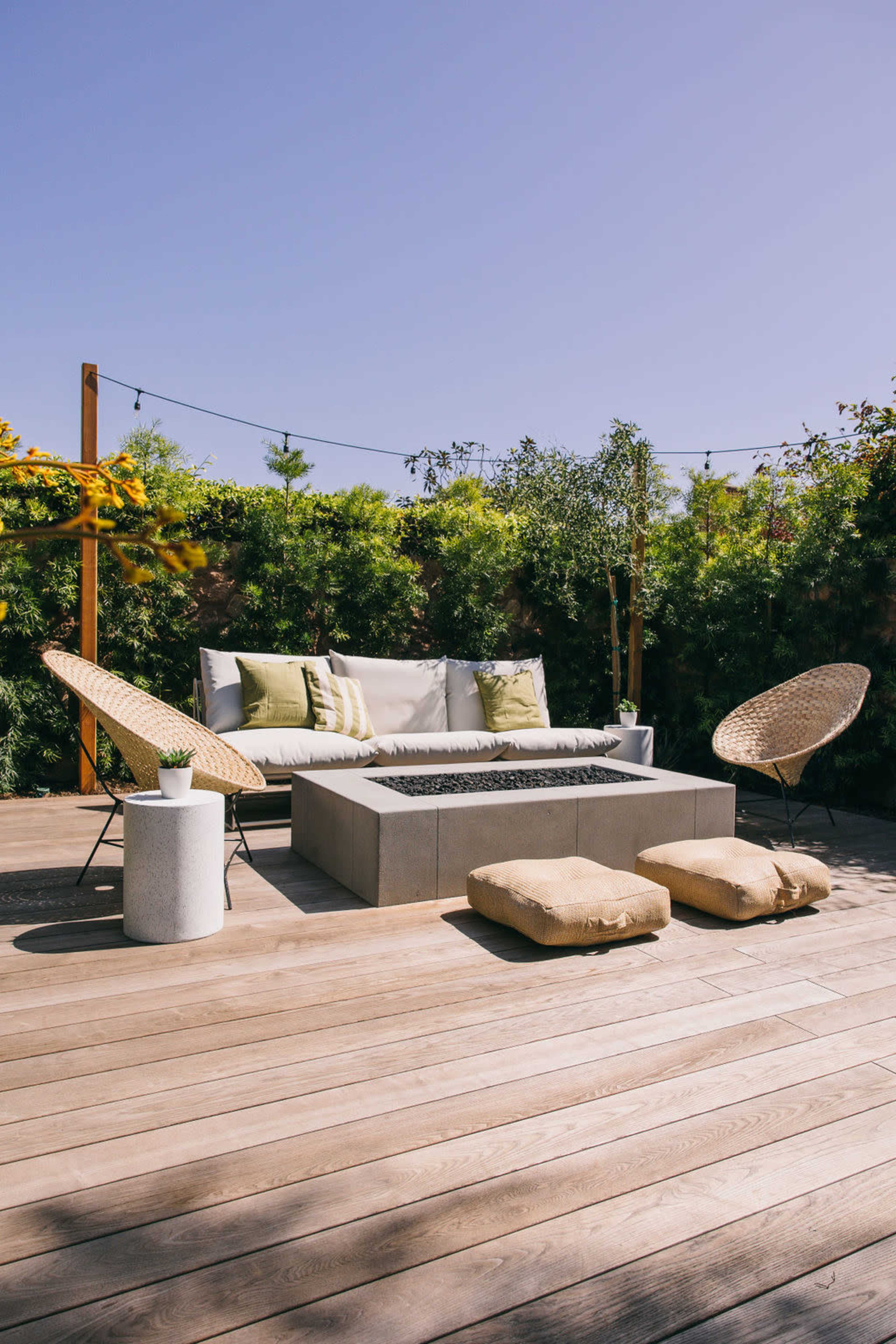 A stylish outdoor seating area with a fire pit, surrounded by greenery and featuring comfortable lounge chairs and cushions.