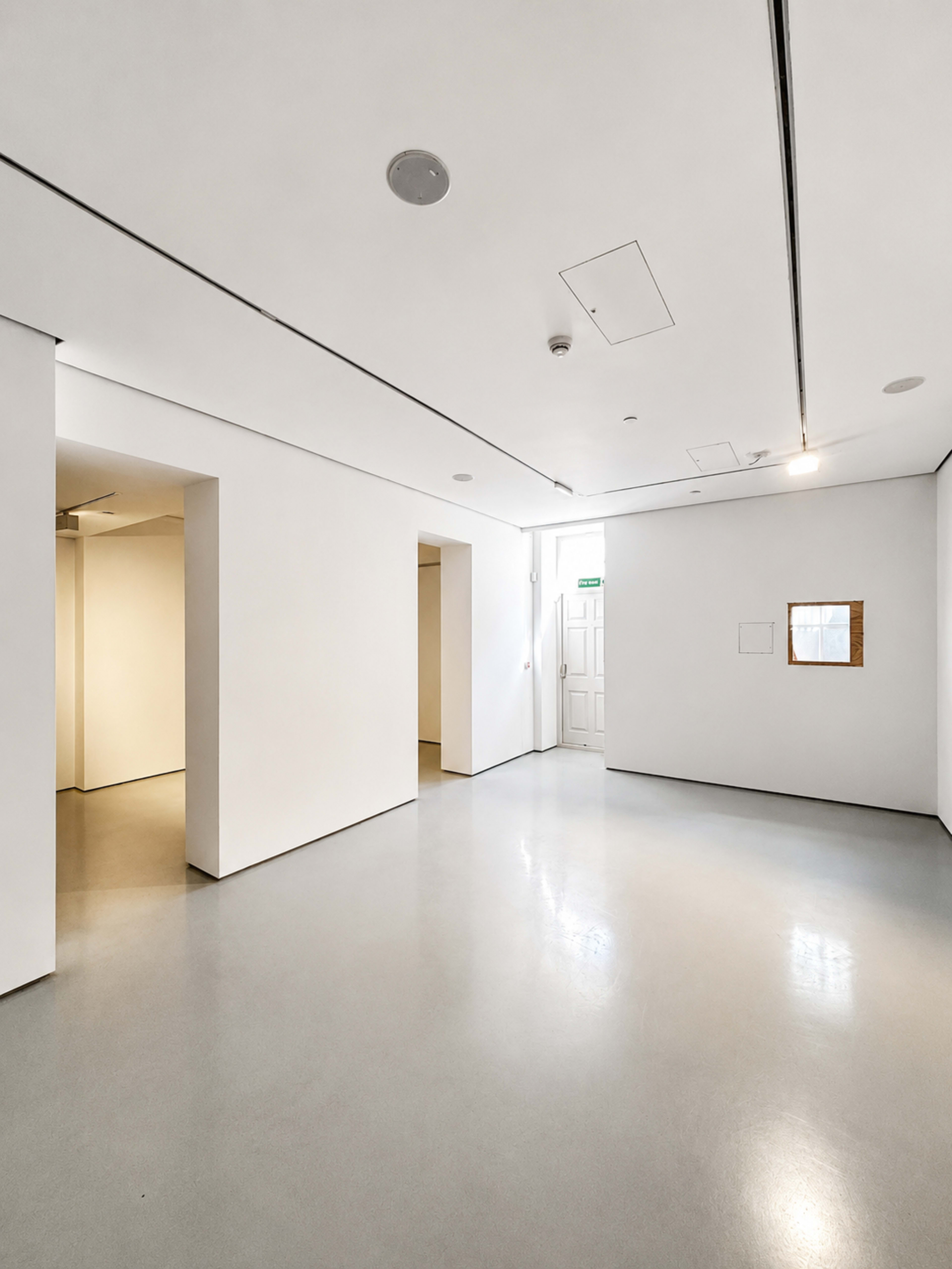 The image shows a minimalist, white-walled gallery space with a polished gray floor and two openings leading to other areas.