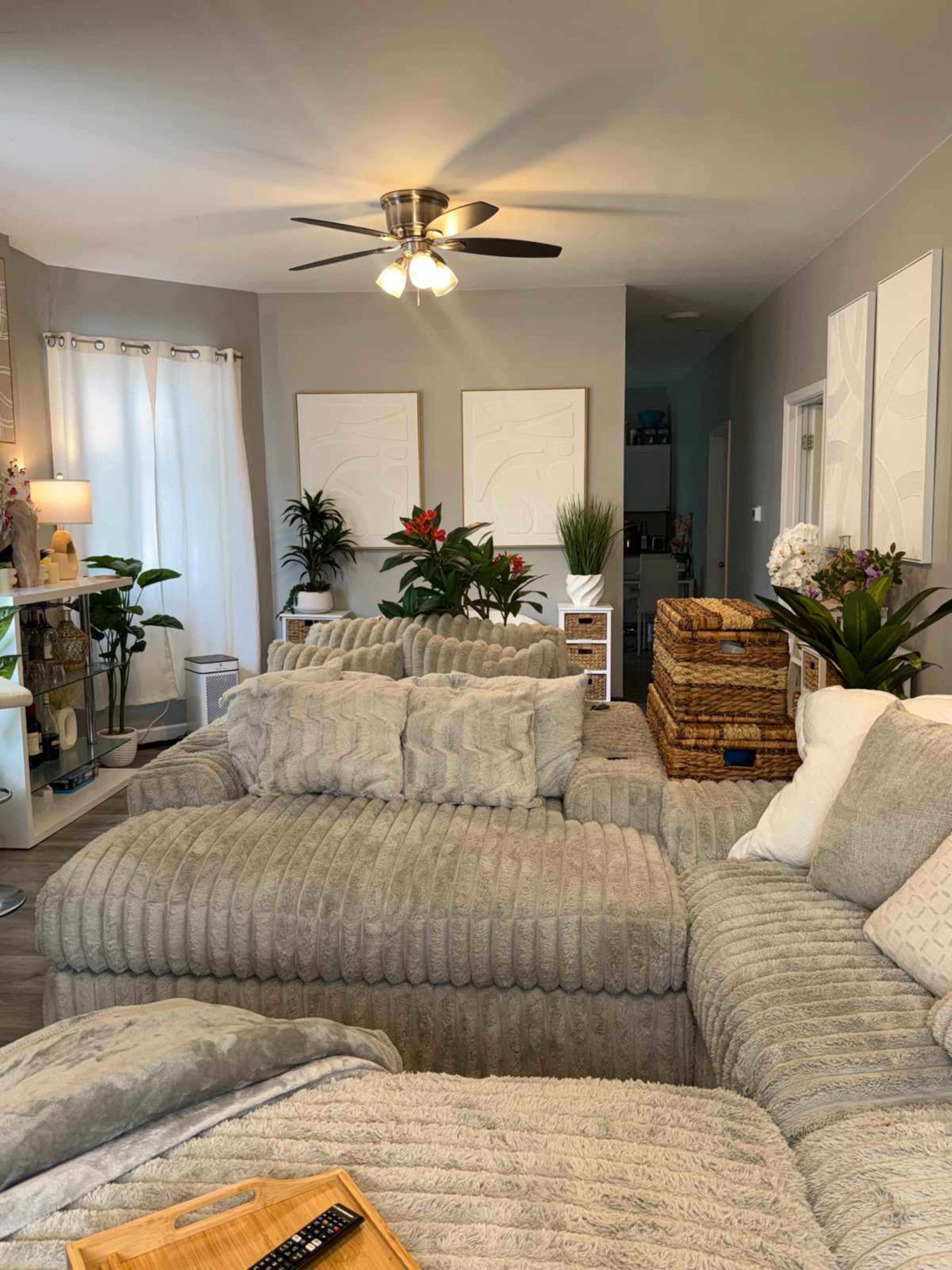 The image shows a cozy living room featuring a large, textured sectional sofa, indoor plants, decorative wall art, and a ceiling fan.