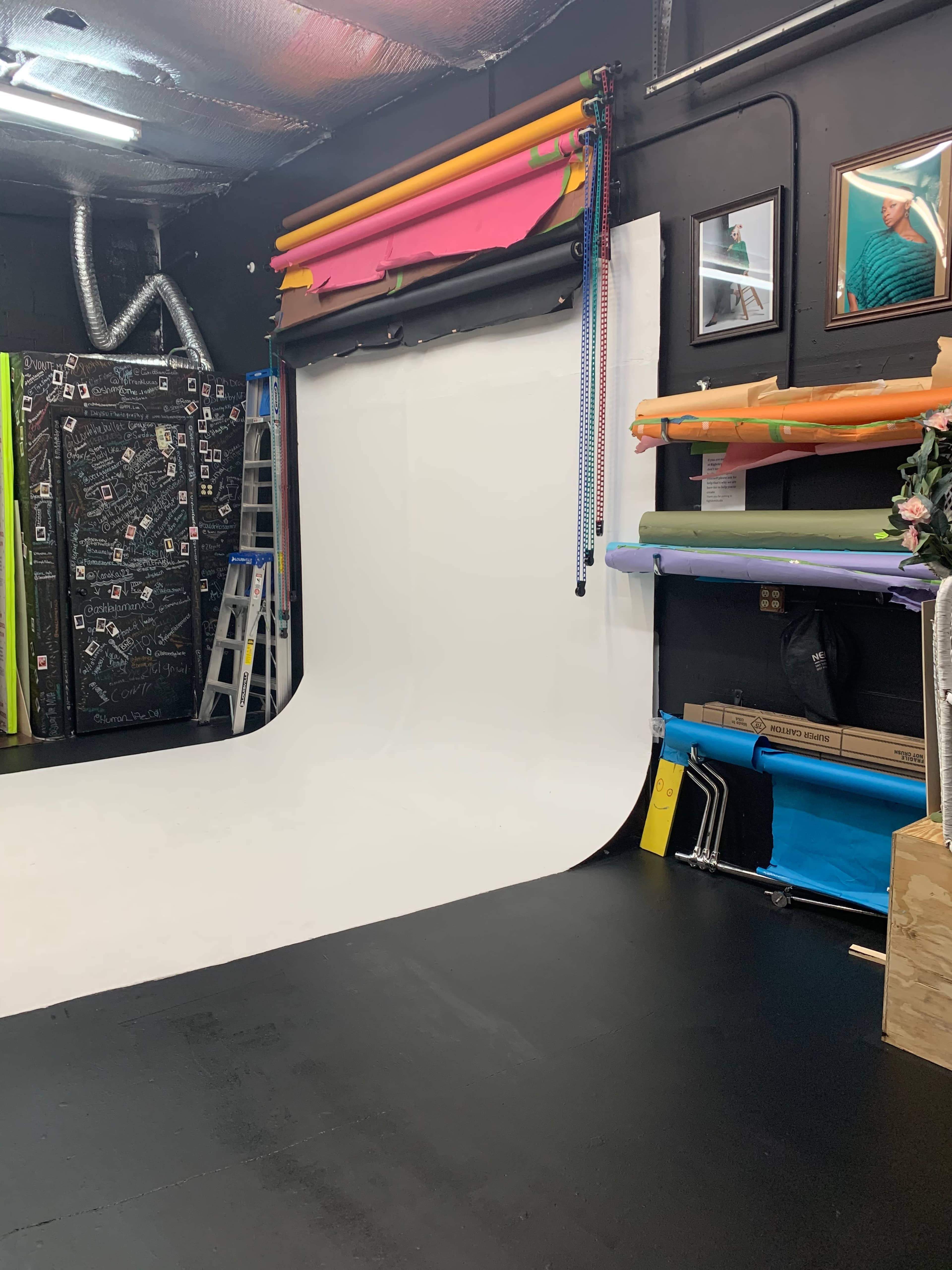 The image shows a photography studio with a white backdrop, colorful rolls of seamless paper, and various props organized on the walls and floor.