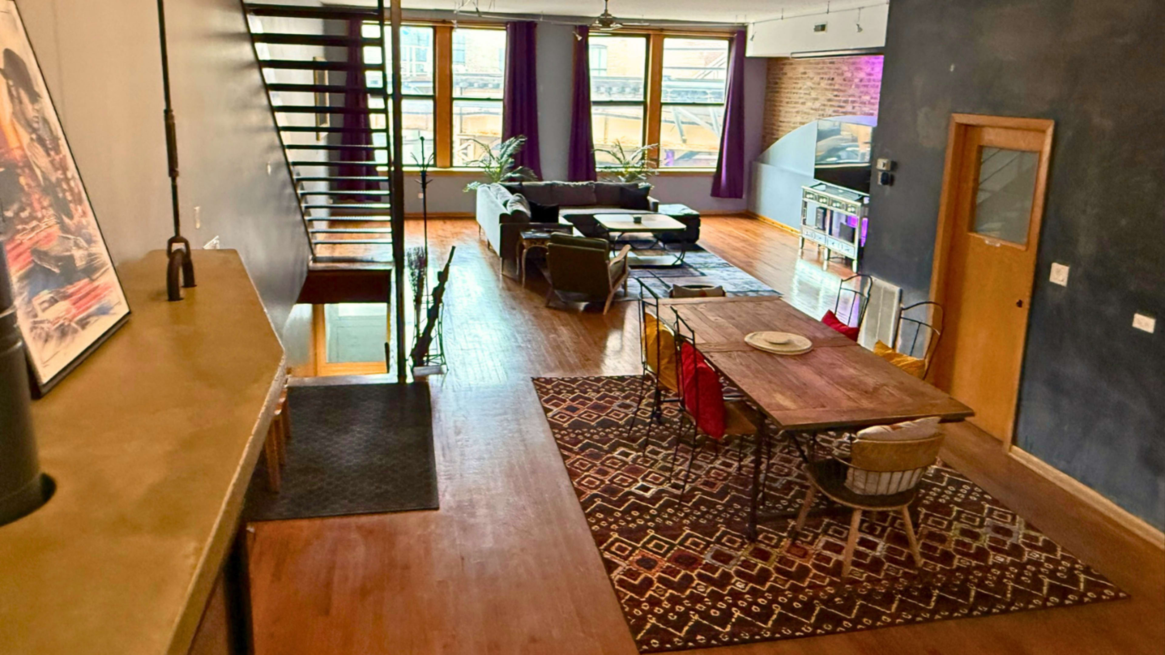 The image shows an open-concept living space featuring a dining area with a table and chairs, a staircase, and a lounge area with a sectional sofa and plants.