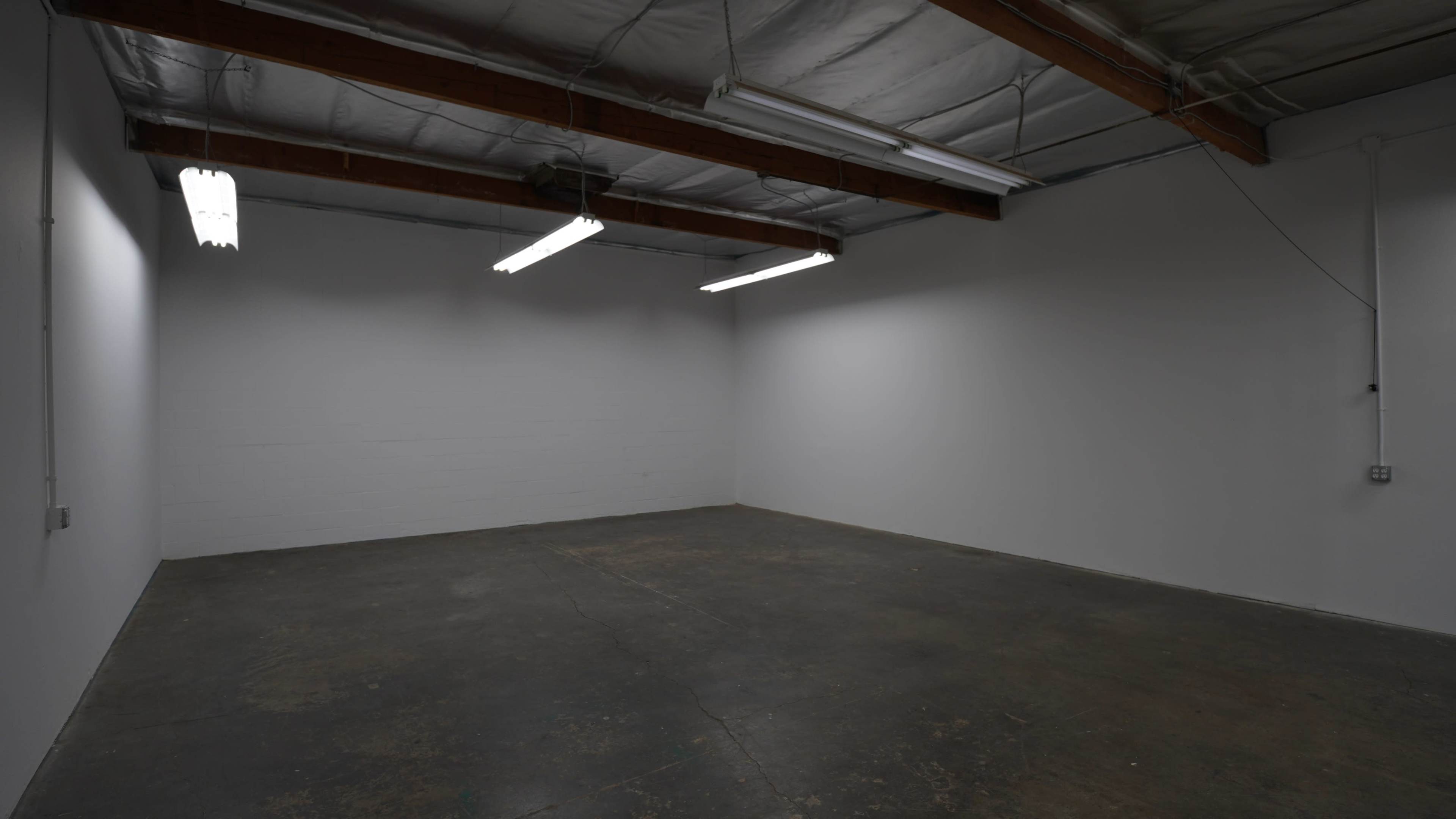 The image shows an empty room with bare concrete flooring and plain white walls, illuminated by fluorescent lights hanging from a ceiling.