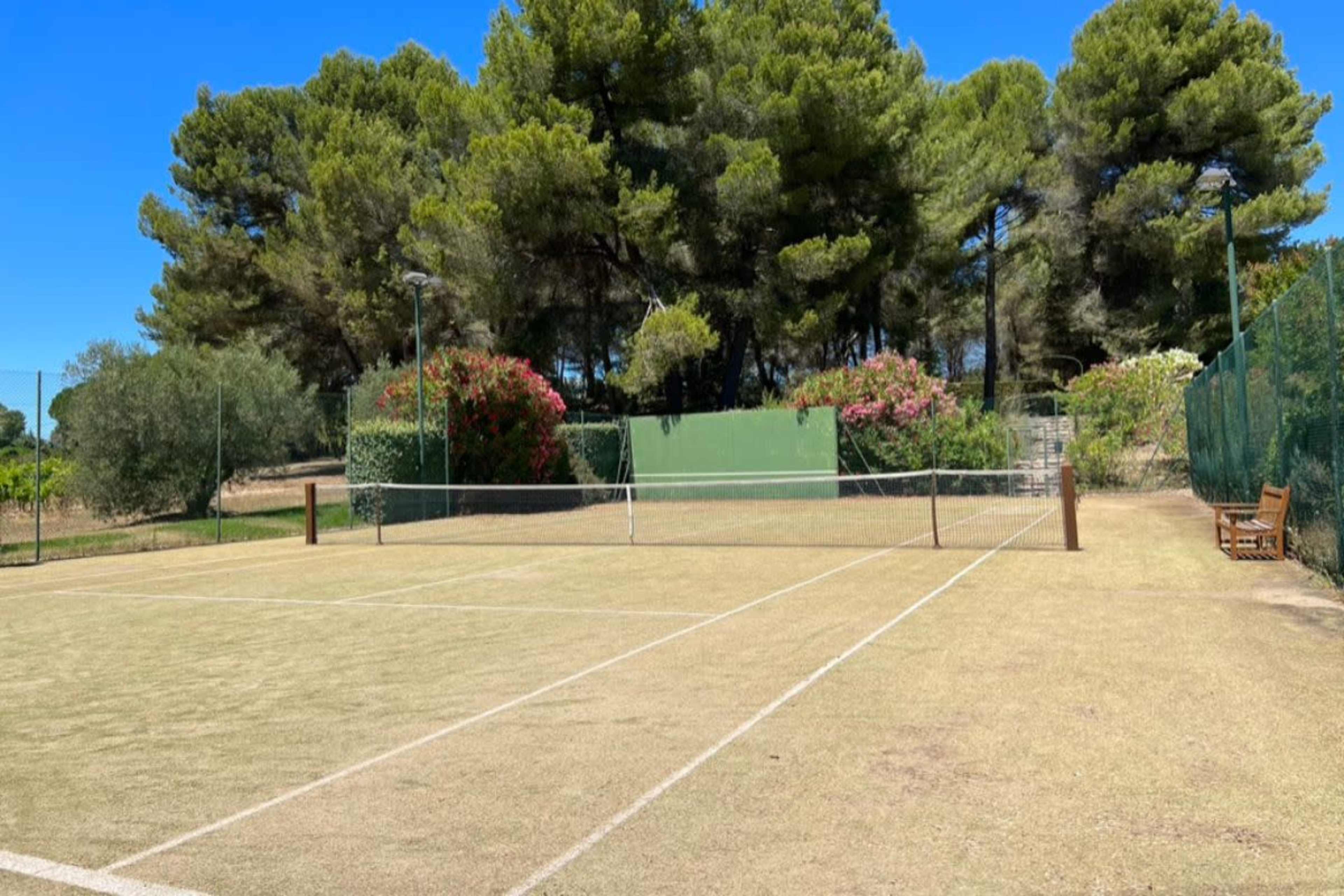 A tennis court with a sandy surface and green fencing is surrounded by tall trees and flowering shrubs.