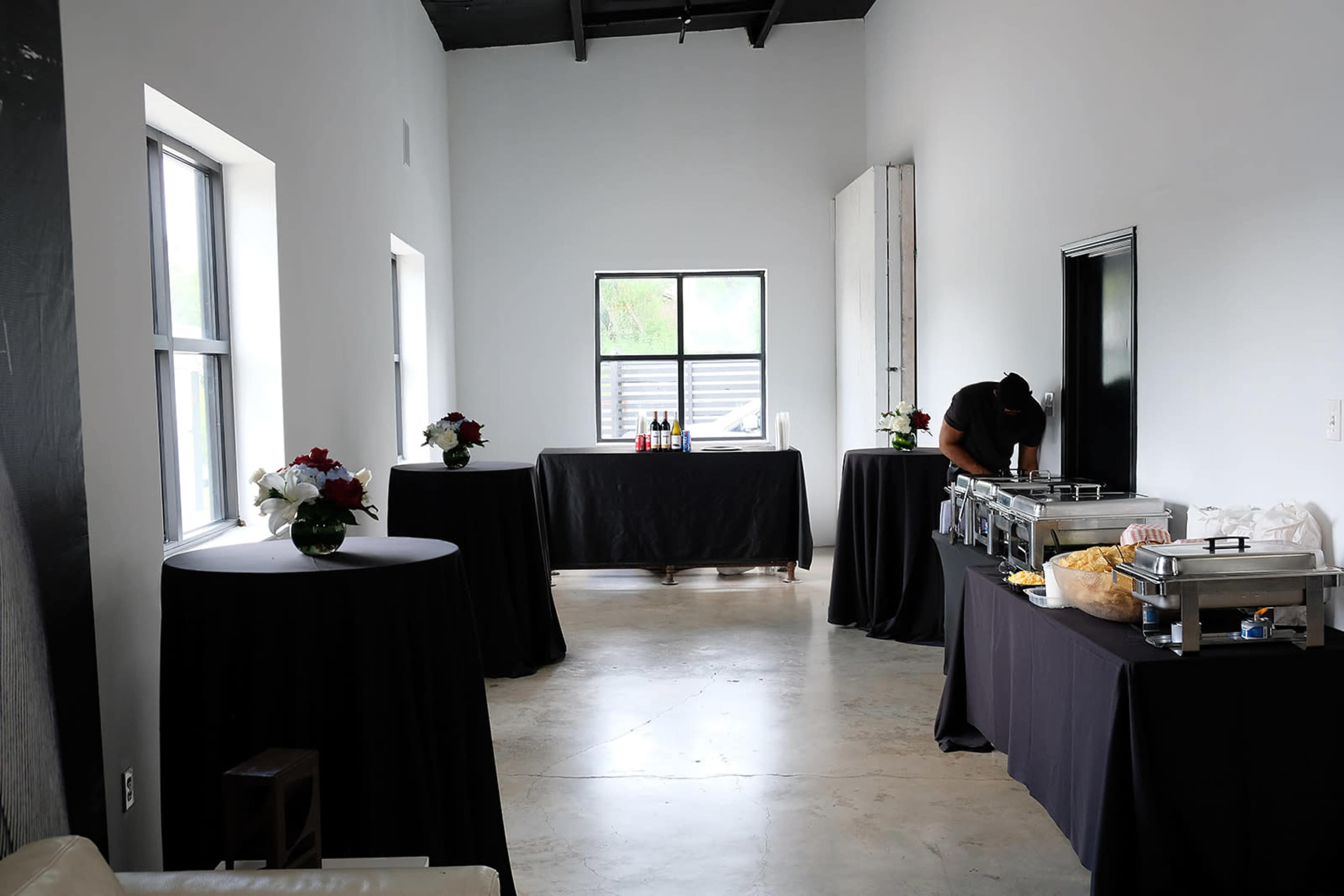 A spacious room with two windows, decorated tables with black tablecloths, and a buffet setup on one side.