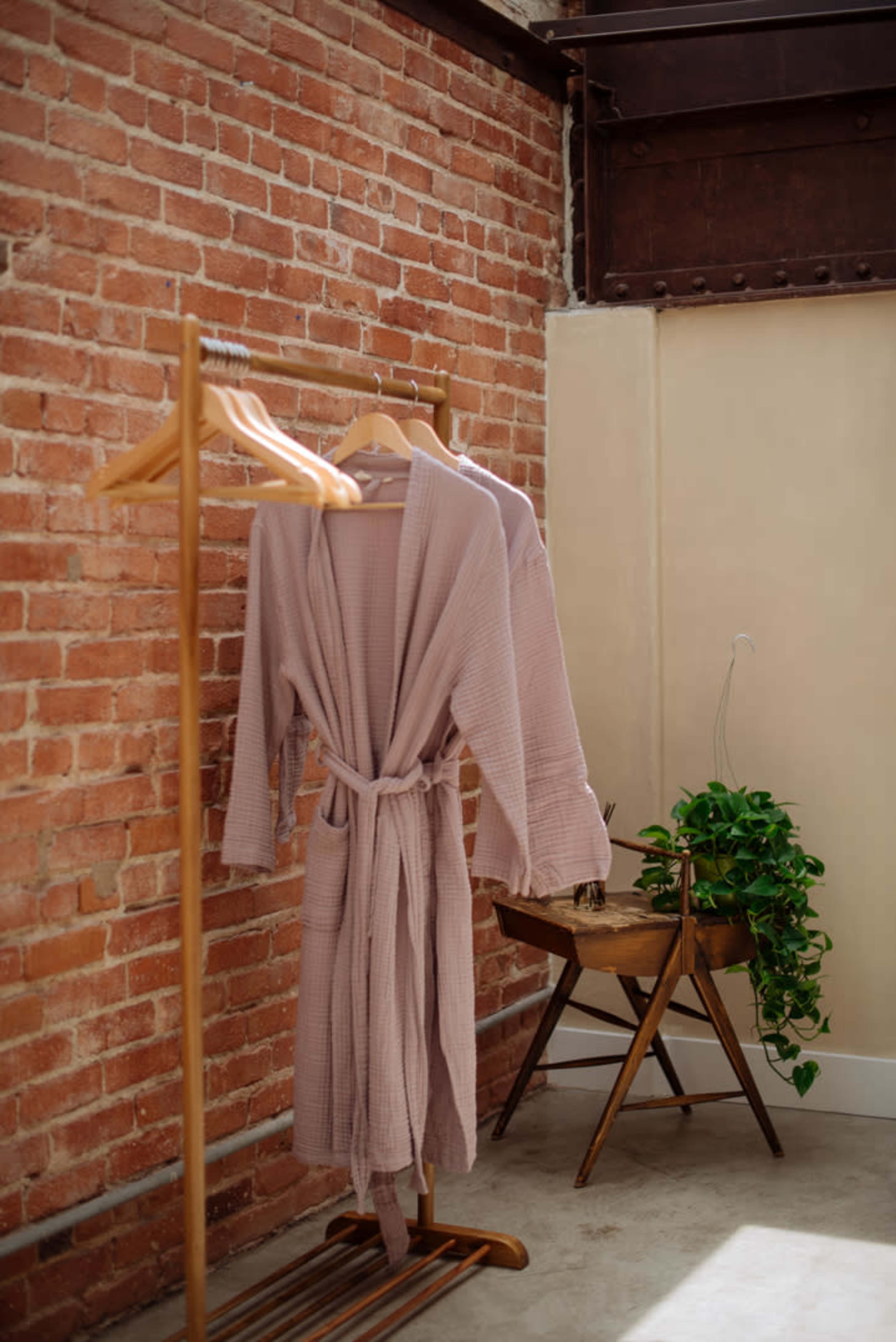 A pair of light-colored bathrobes hangs on a wooden rack next to a potted plant in a room with exposed brick walls.