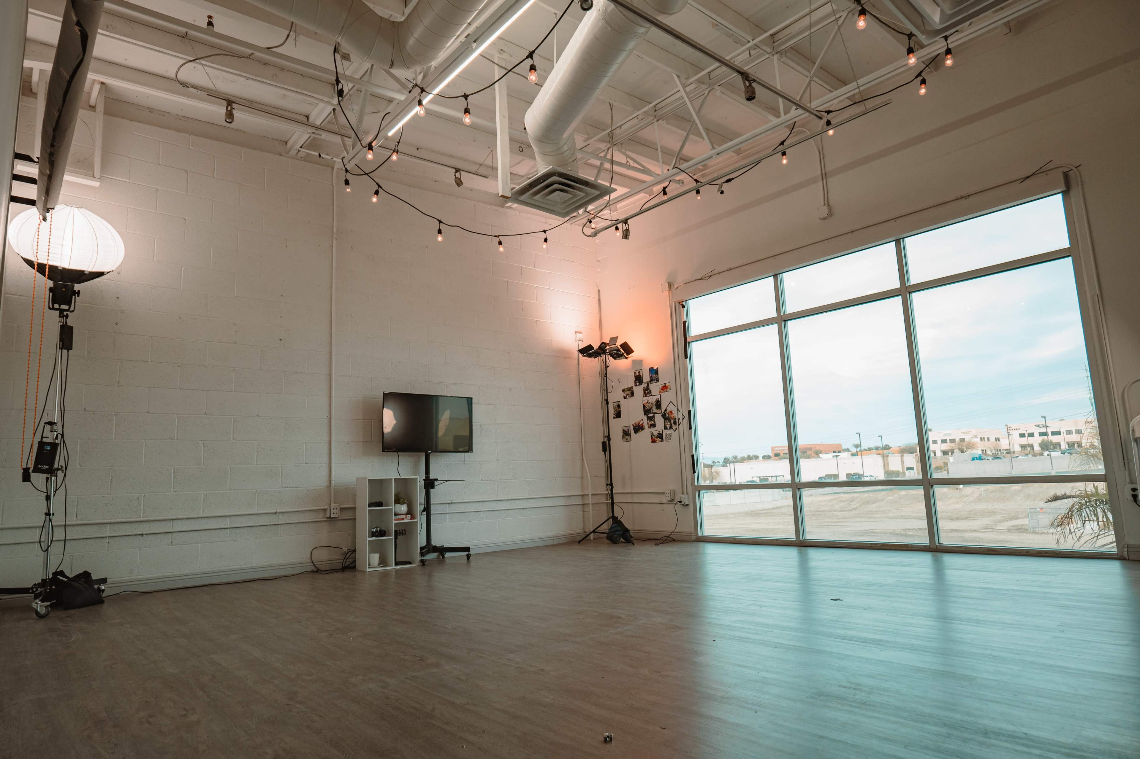 An empty room features a large window with natural light, a television mounted on the wall, and decorative string lights overhead.