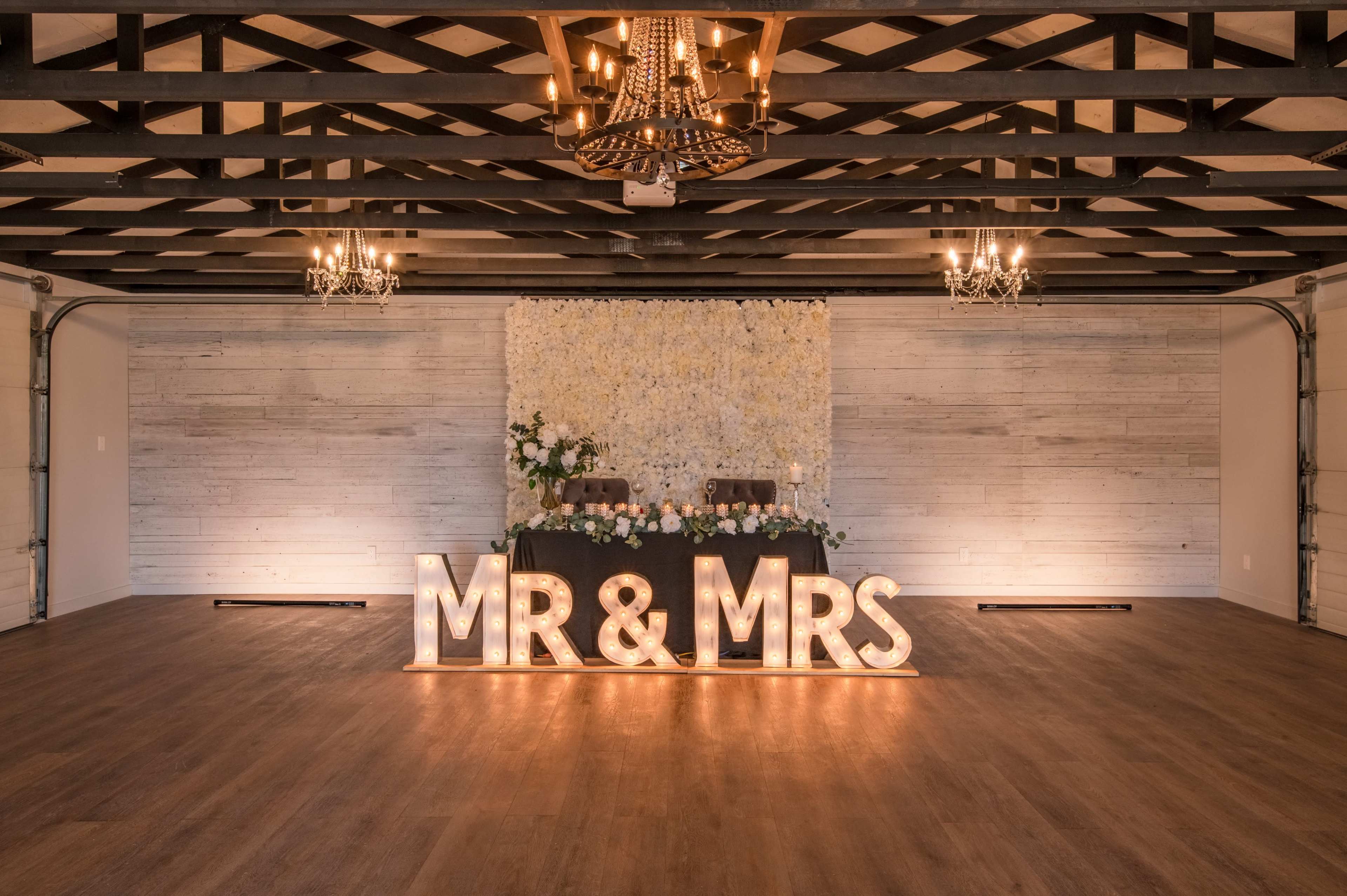 The image shows a decorated reception space featuring a "MR & MRS" light-up sign in front of a floral backdrop and a head table.