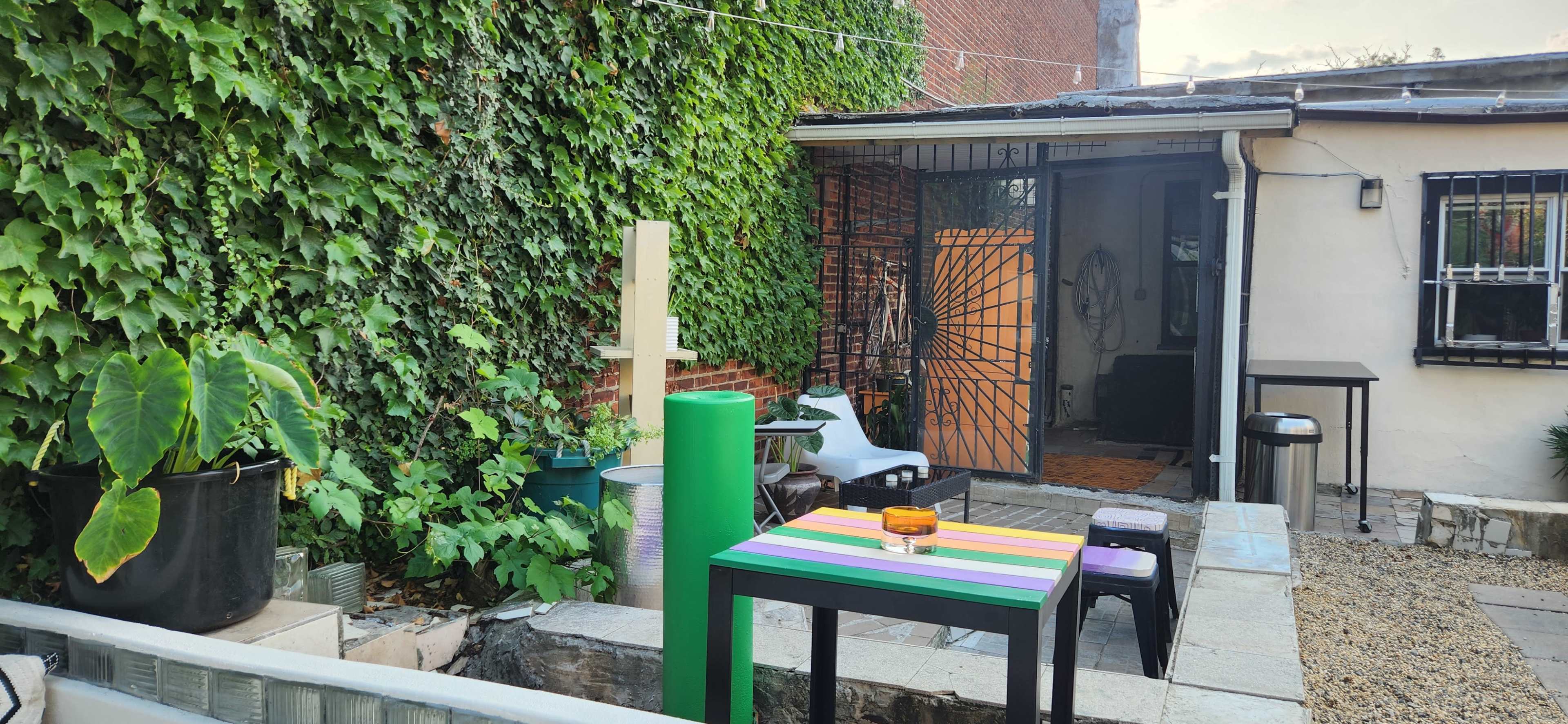 The image shows a small outdoor area with a multi-colored table, various plants, and a building with a gated entrance.