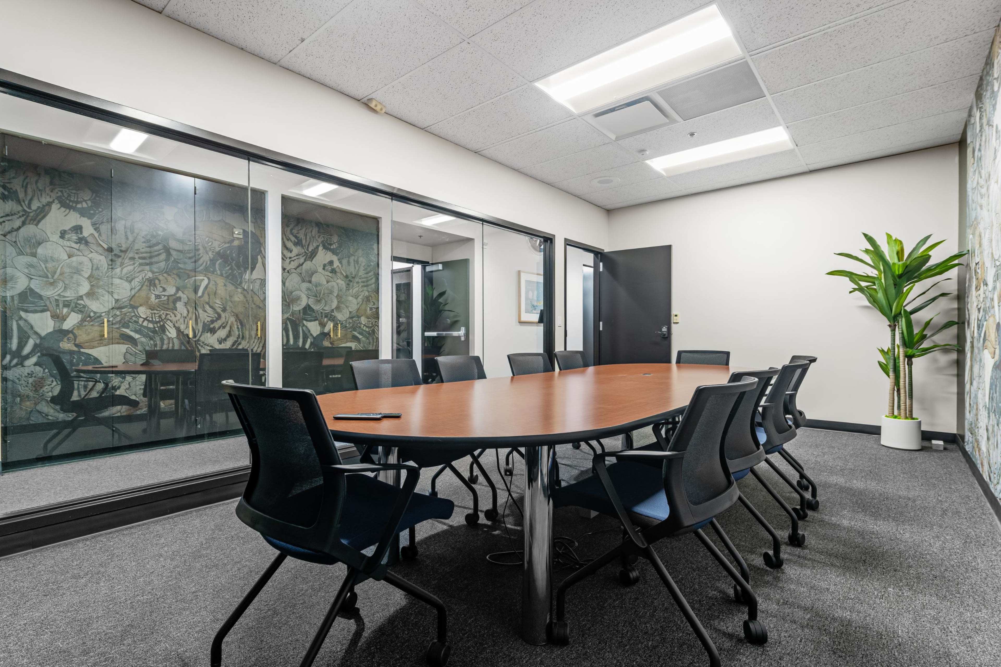 The image shows a conference room featuring a large round table surrounded by black chairs, with glass walls and a plant in the corner.