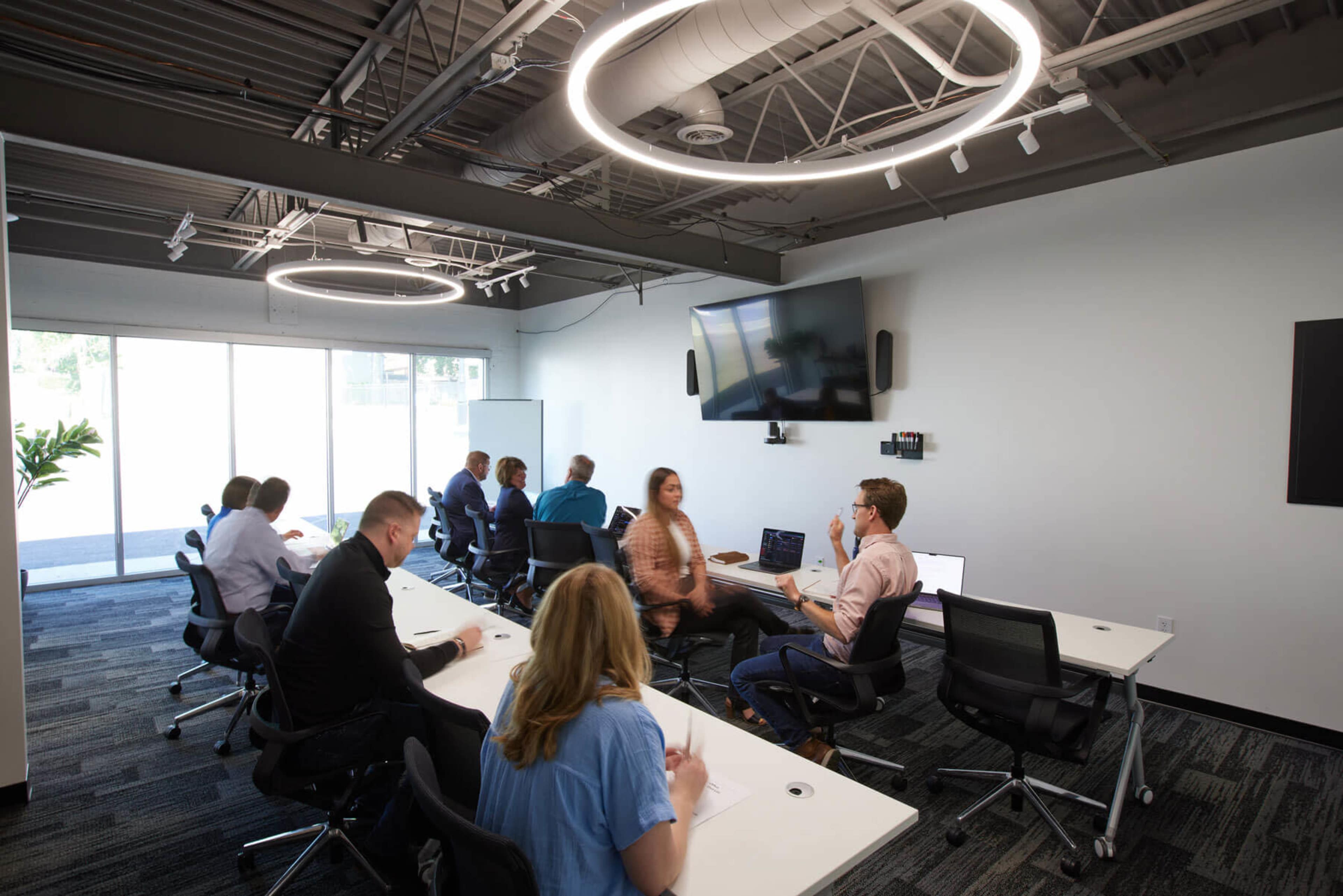 A group of people is seated at tables in a modern conference room, engaging in discussions while a presentation takes place on a screen.