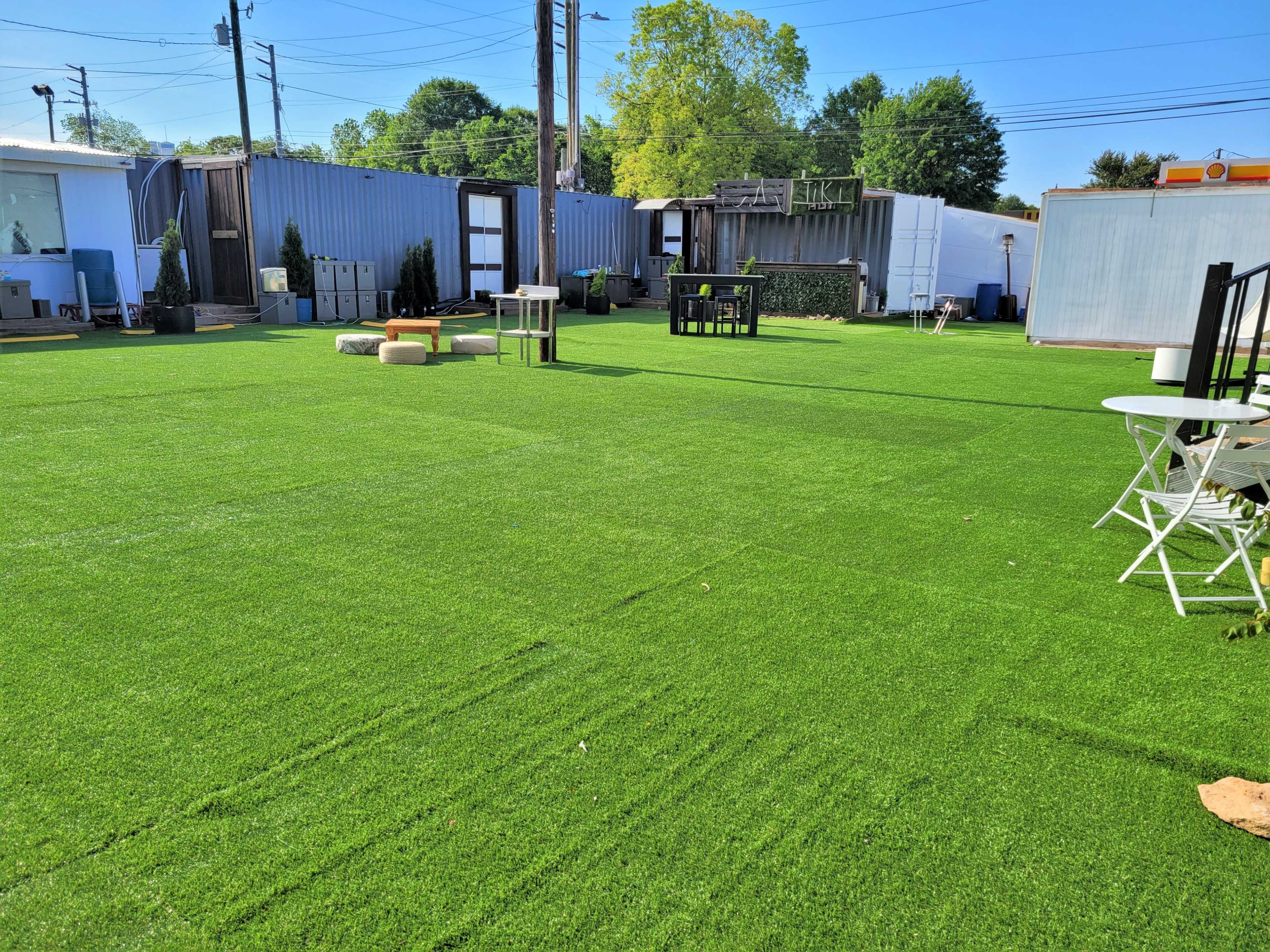 The image shows a spacious outdoor area with artificial grass, surrounded by various structures and furniture.