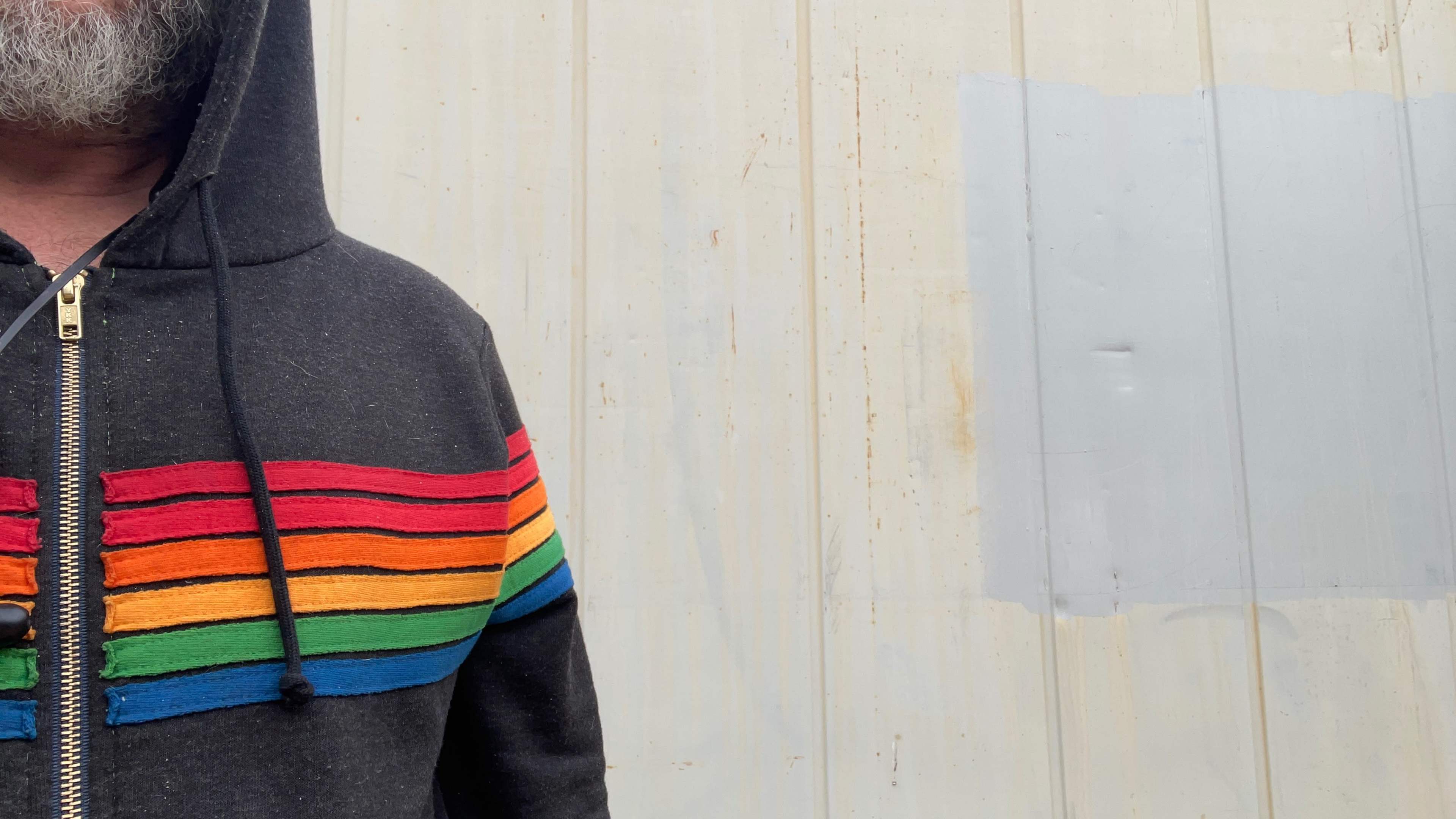 A man wearing a hoodie with rainbow stripes stands next to a textured, light-colored wall.
