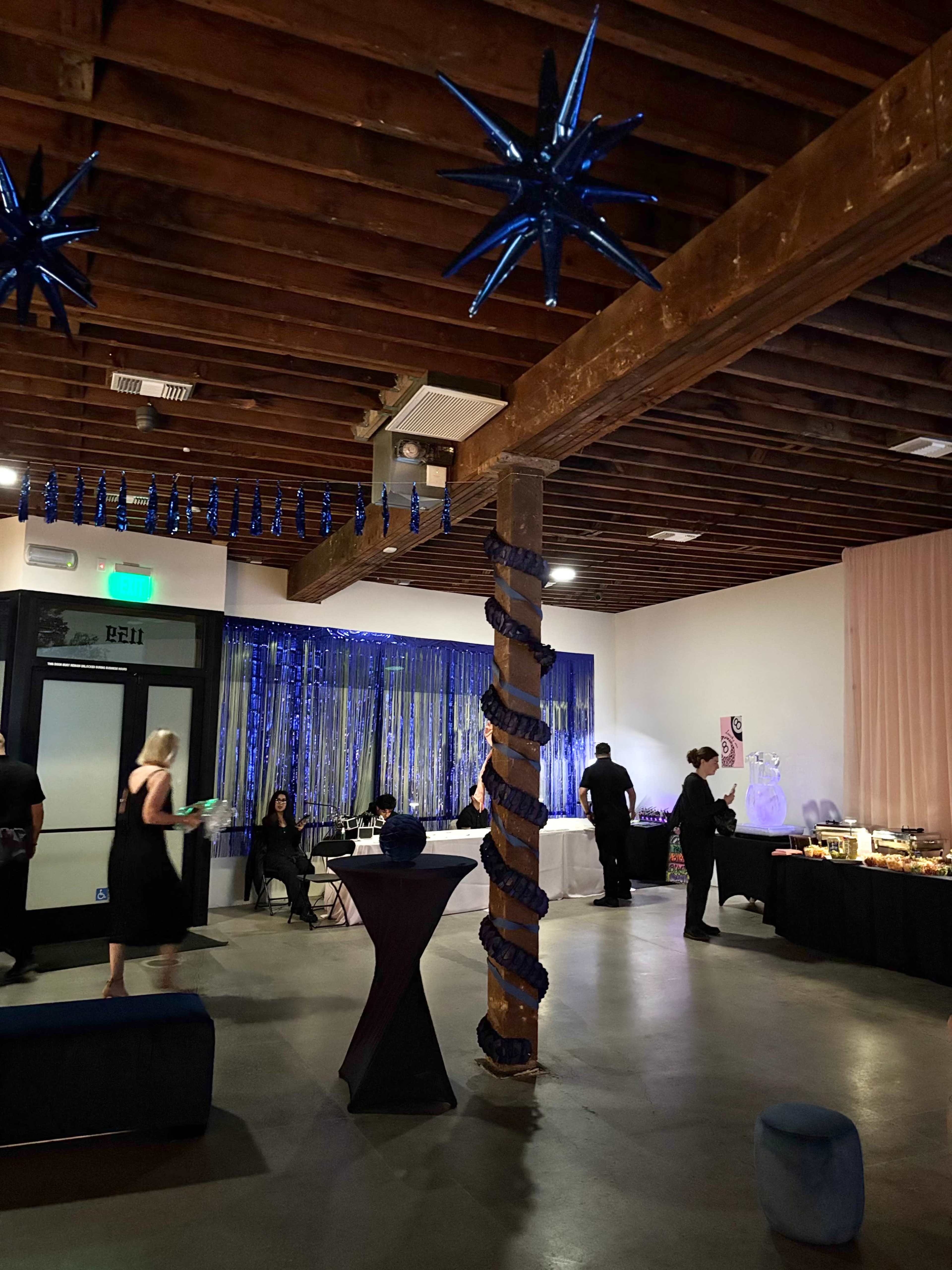 The image shows a decorated event space with blue star-shaped ornaments hanging from the ceiling, a dining area with tables, and people mingling.