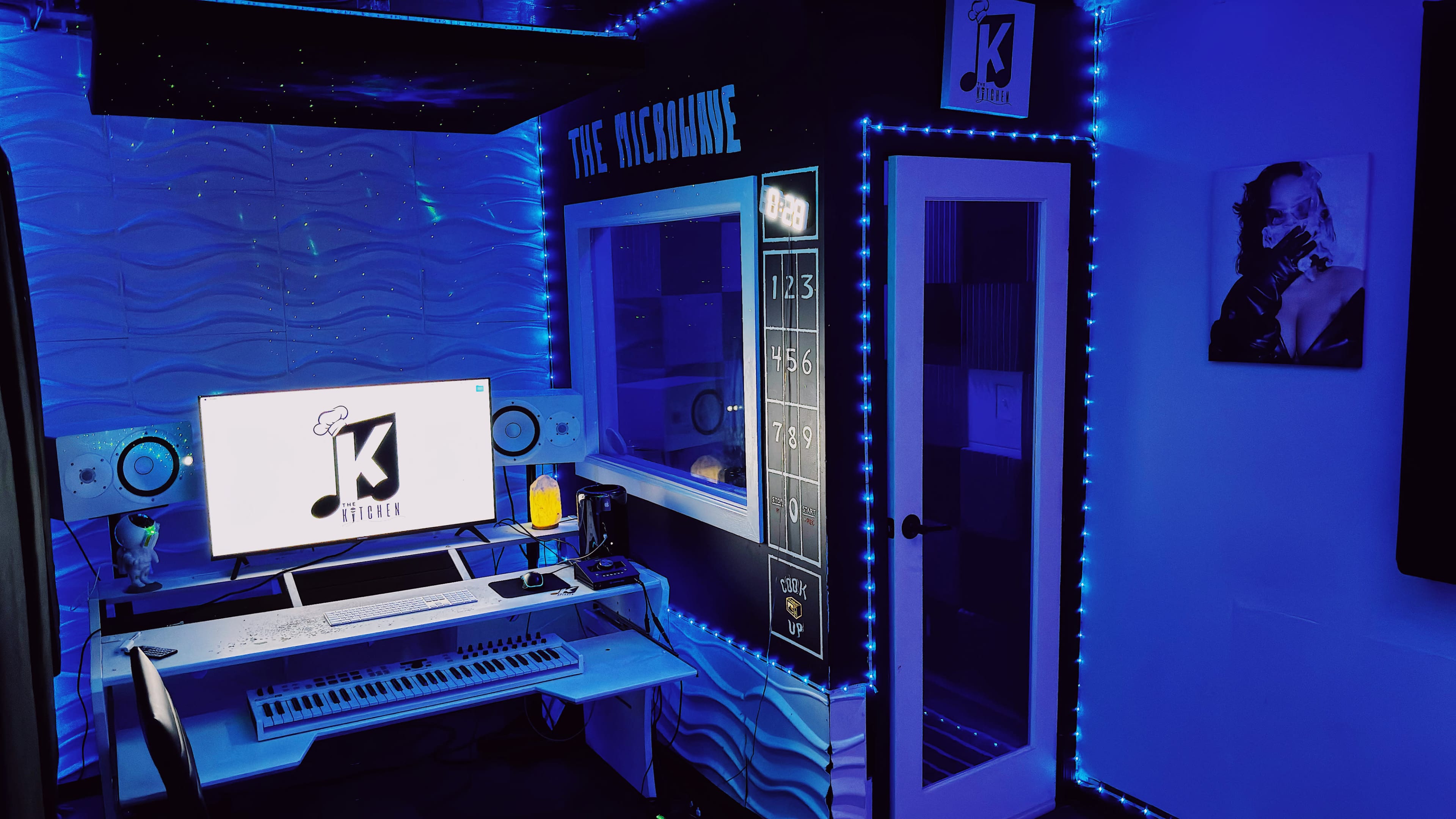 The image depicts a music recording studio featuring a keyboard, a computer setup, and illuminated walls with blue LED lights.