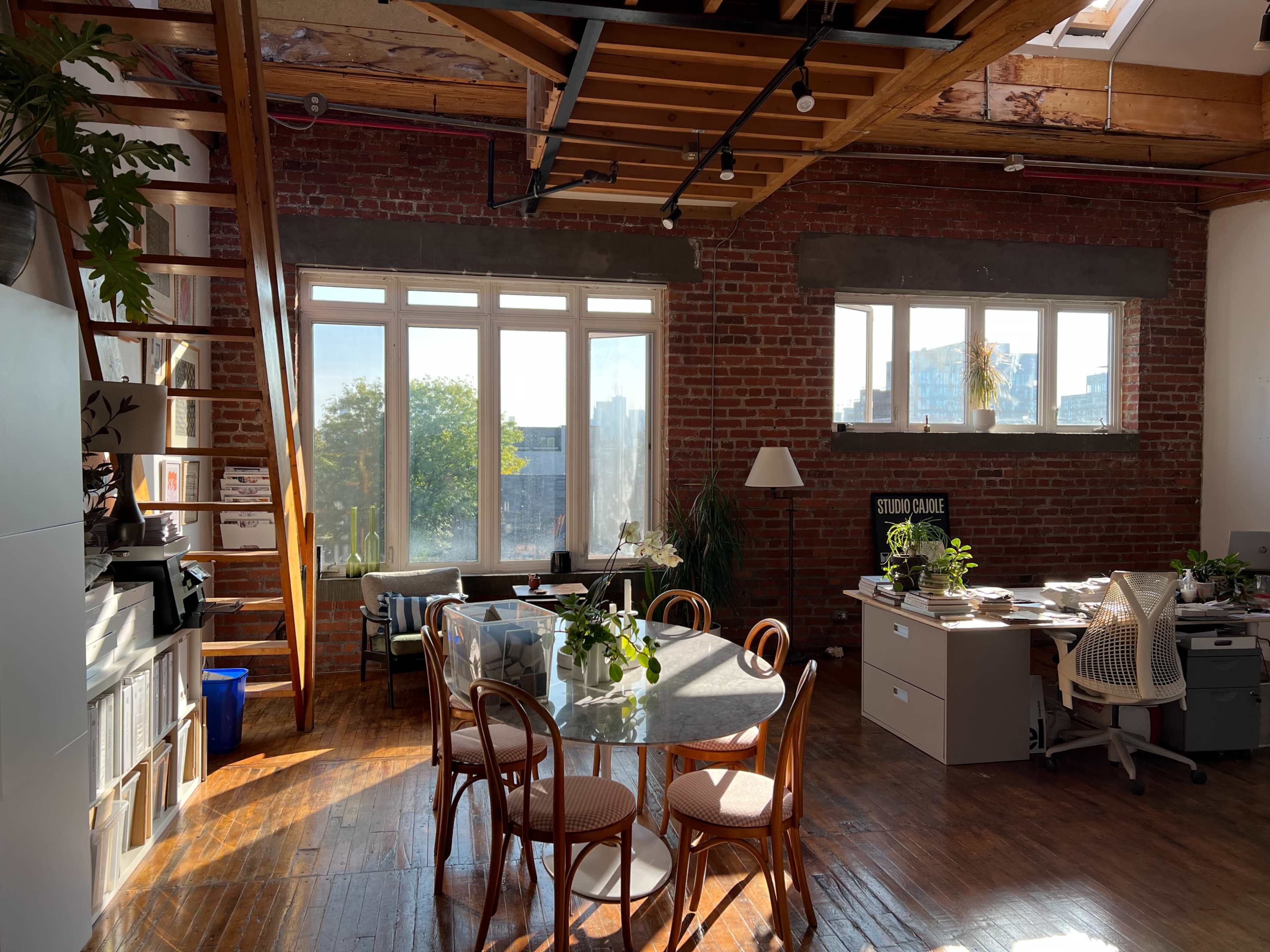 A spacious office with a round table, chairs, and natural light streaming in through large windows, featuring exposed brick walls and wooden beams.