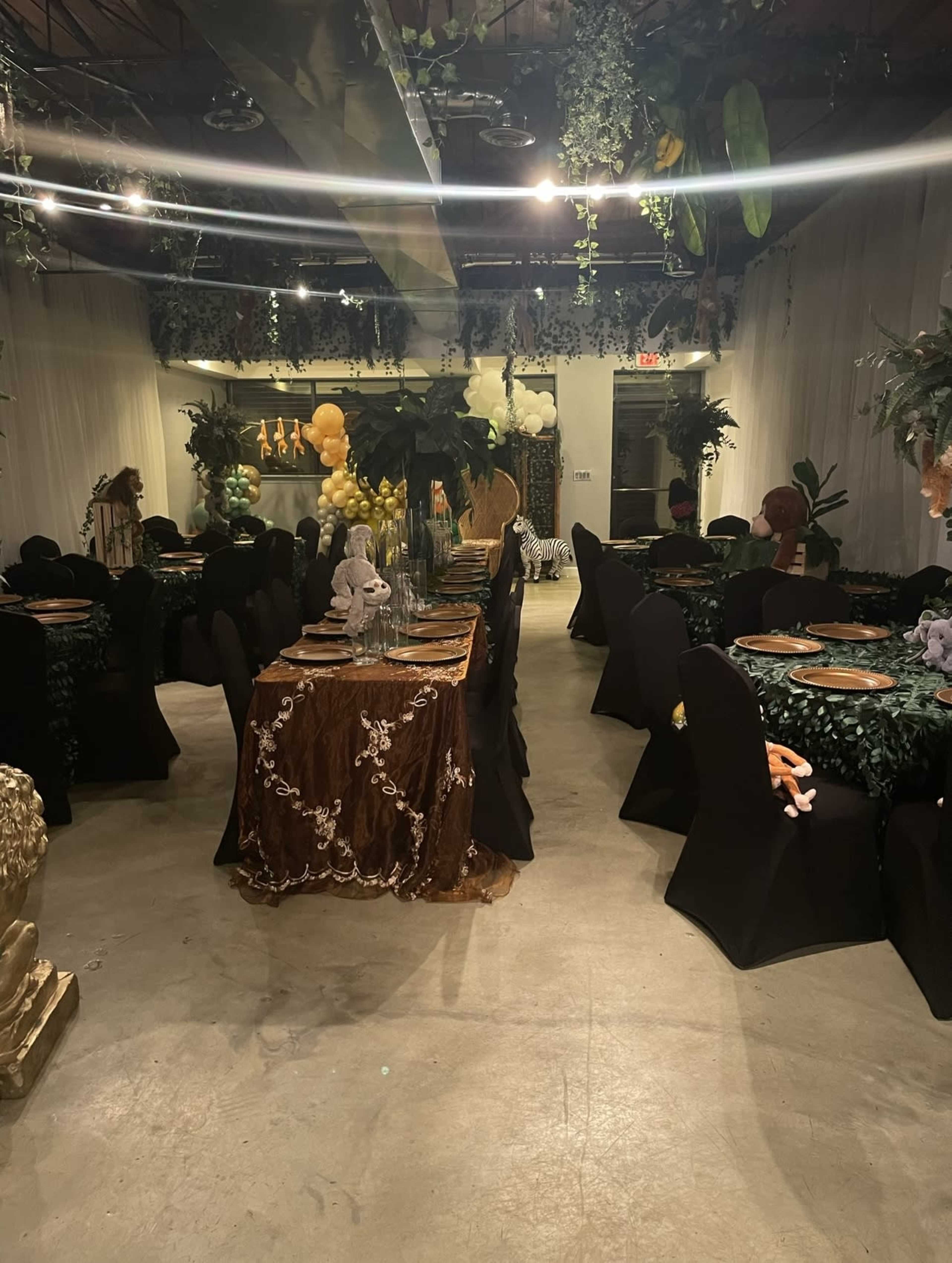 A dining area is set up with black tablecloth-covered tables, adorned with greenery and decorative elements, under bright lights.