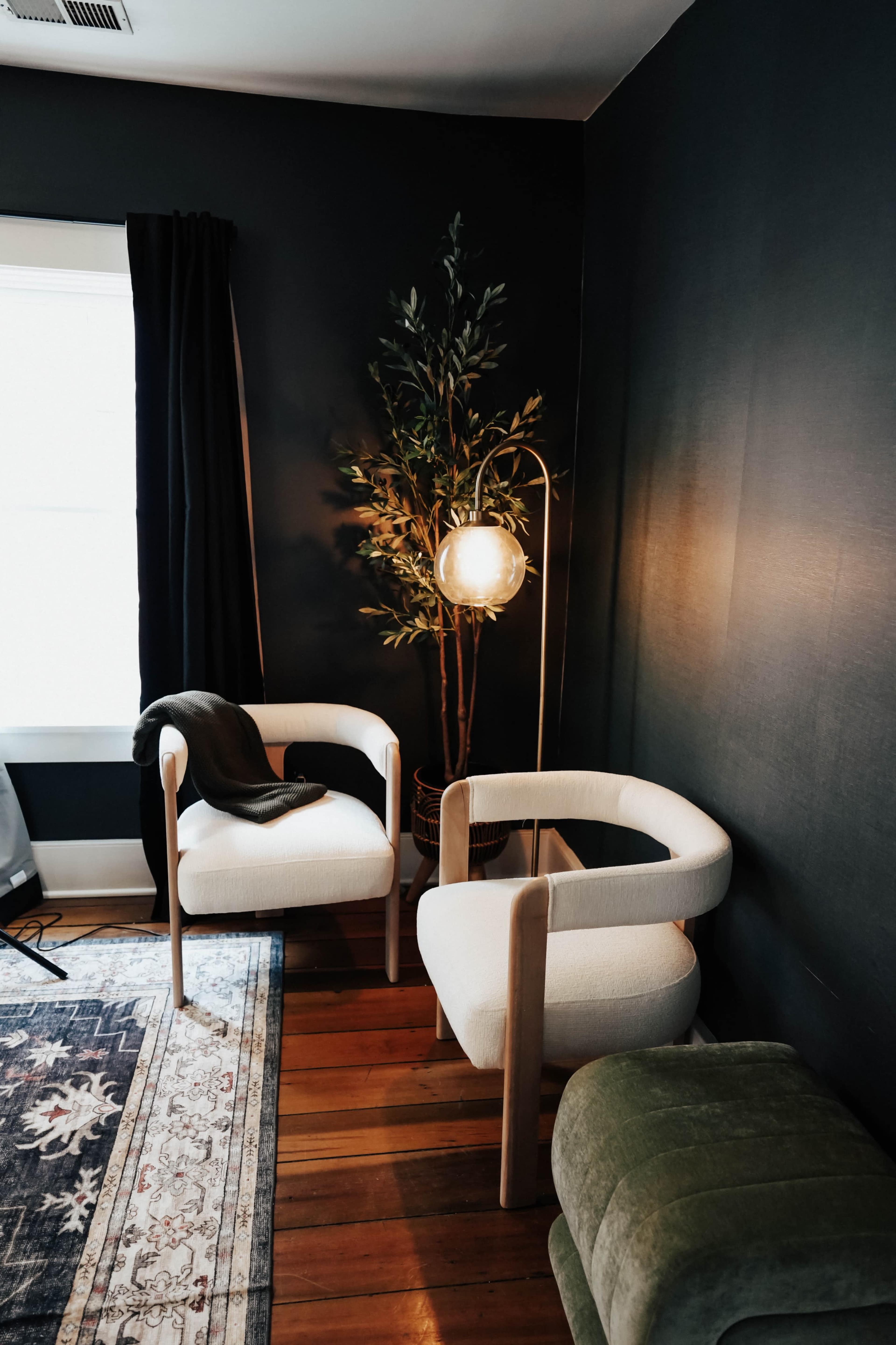 The image shows a corner of a room featuring two white armchairs, a green ottoman, a floor lamp, and a potted plant against dark-painted walls.