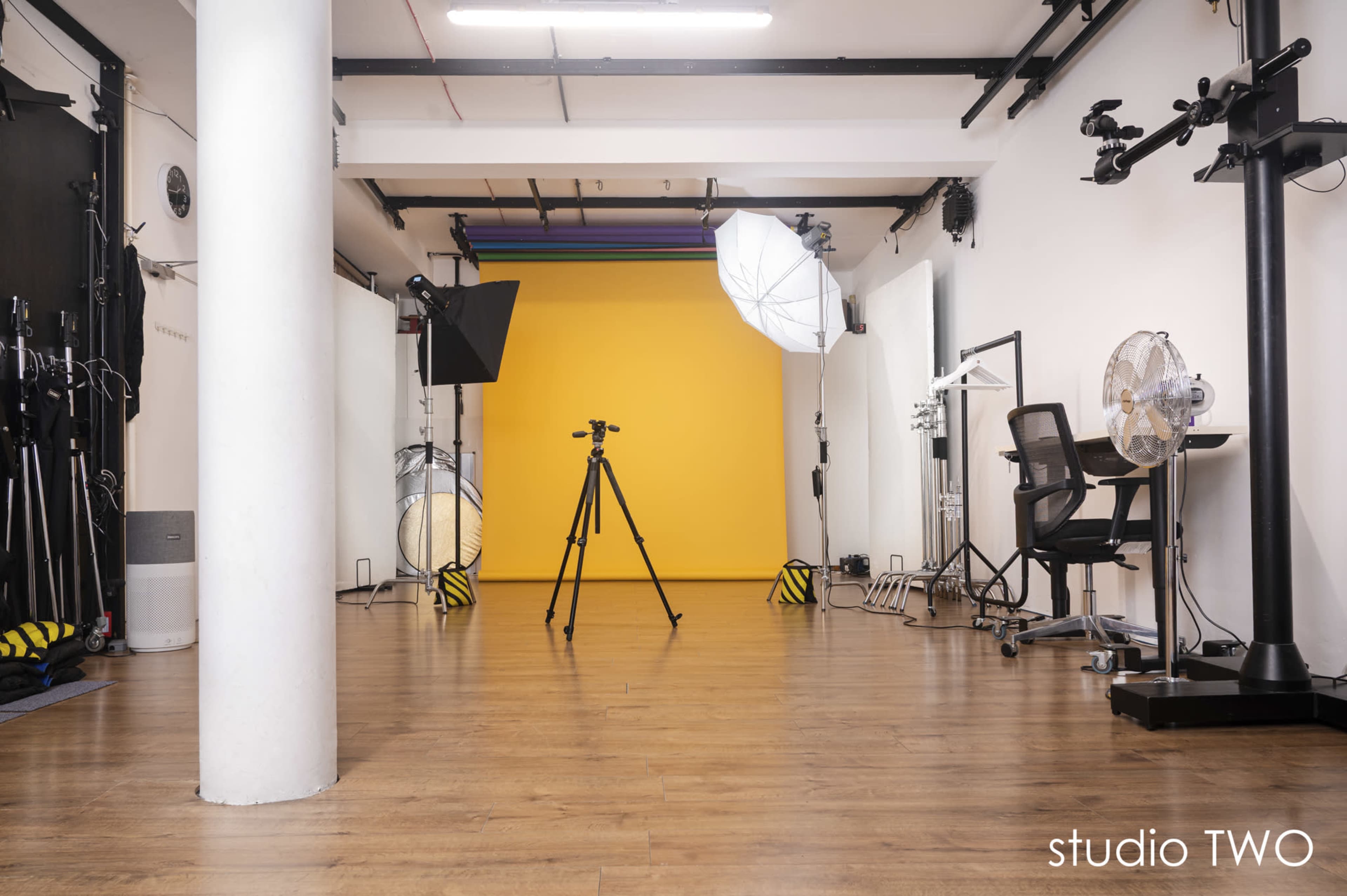 The image shows a well-lit photography studio featuring a yellow backdrop, camera equipment, lighting setups, and a workspace.