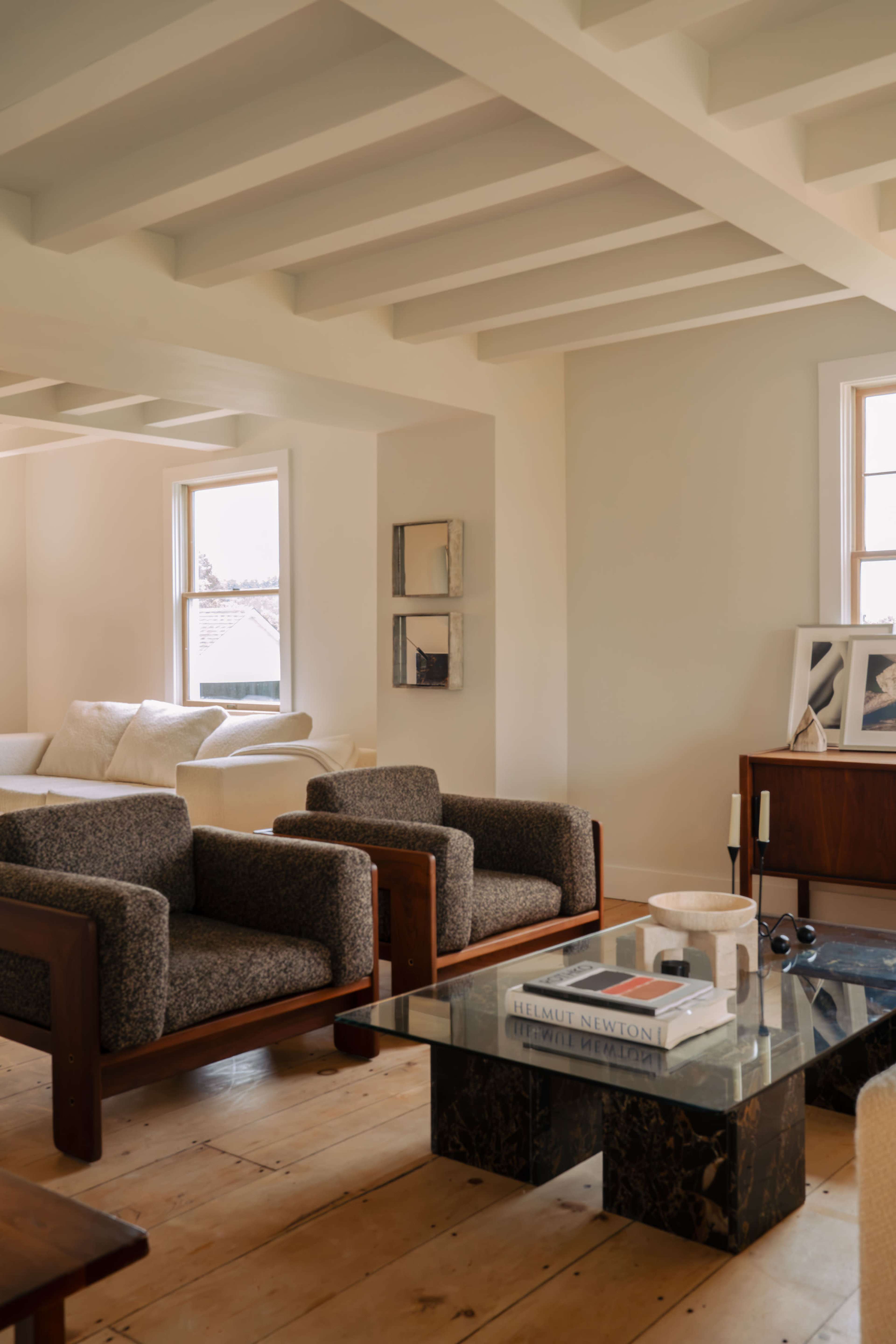 A modern living room featuring two textured armchairs facing a glass coffee table, with a sofa and windows in the background.