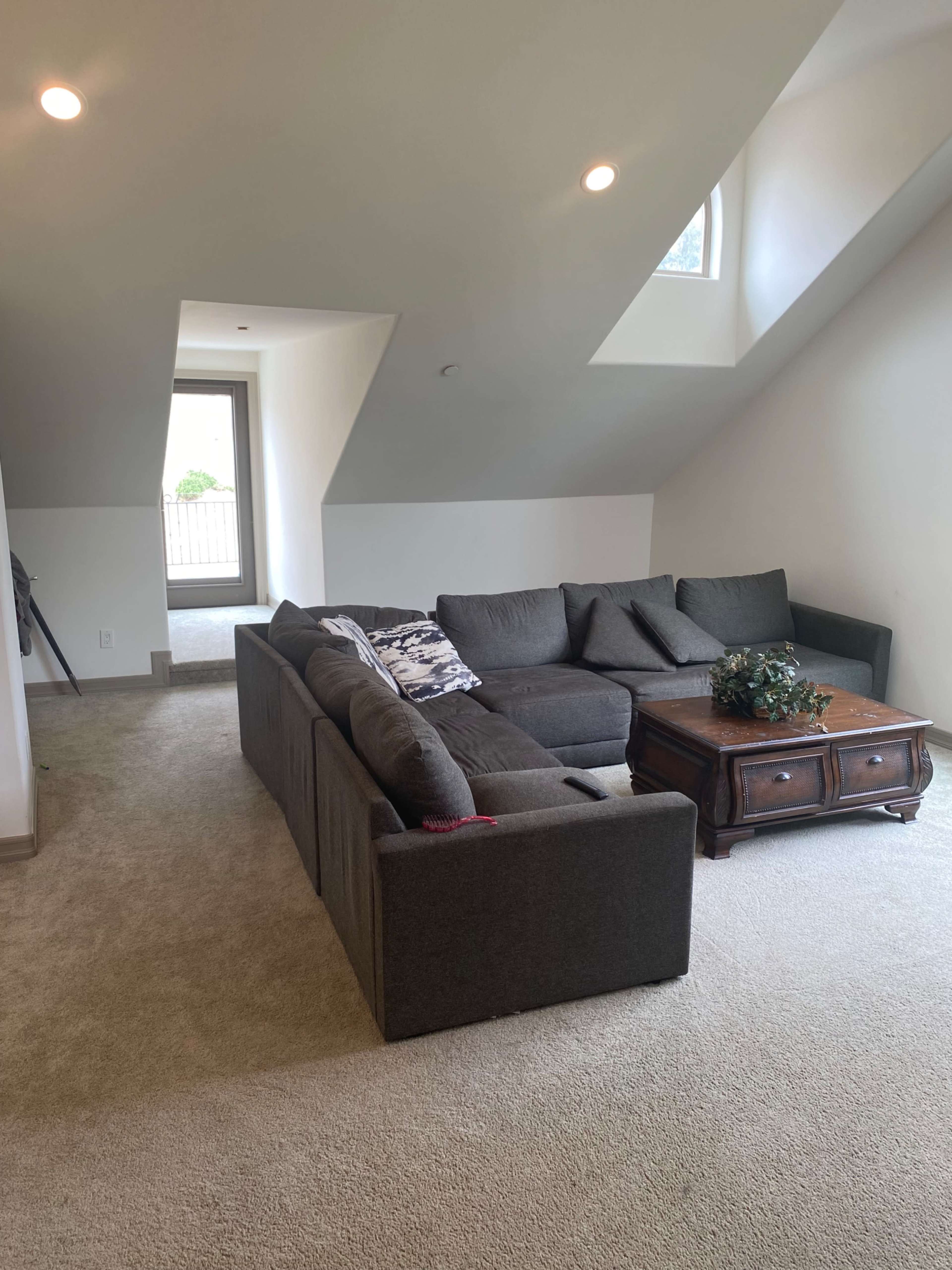 The image shows a living room with a dark gray sectional sofa and a wooden coffee table, situated under a sloped ceiling with a large window in the background.