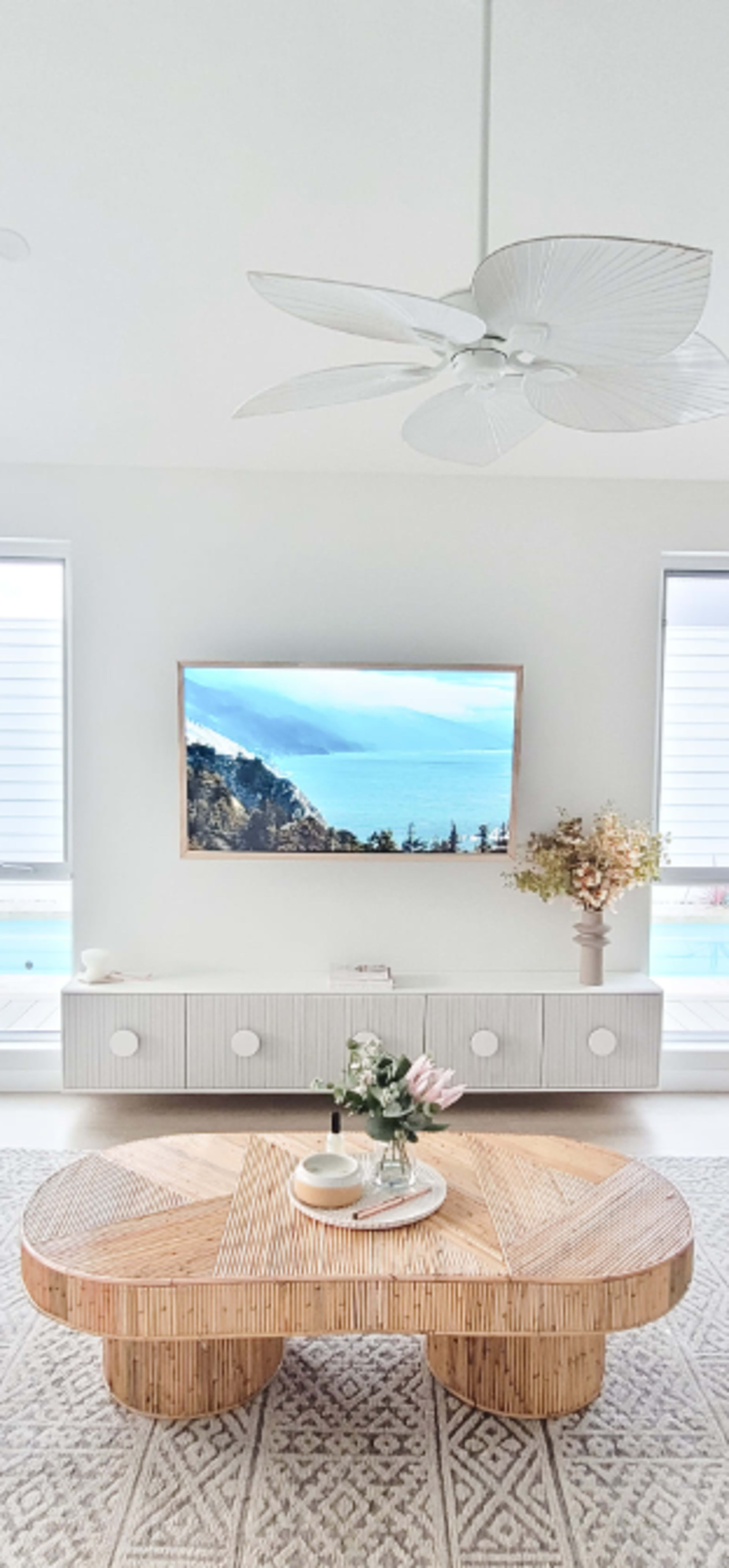 A modern living room features a light-colored wooden coffee table, a flat-screen TV mounted on the wall, and a sideboard with circular handles, accented by natural light from large windows.