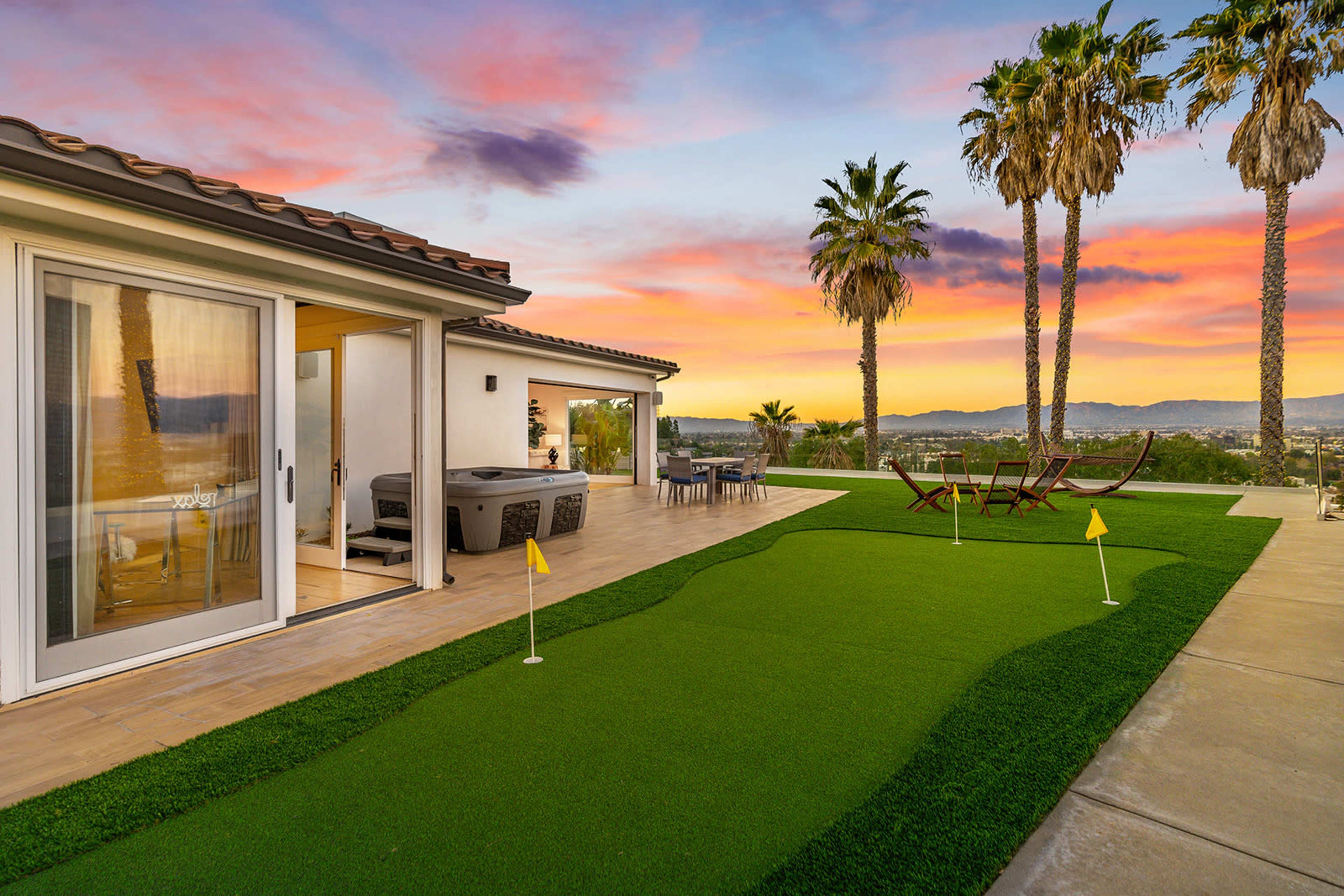 The image shows a modern backyard with a putting green, palm trees, and a view of a sunset sky over rolling hills.