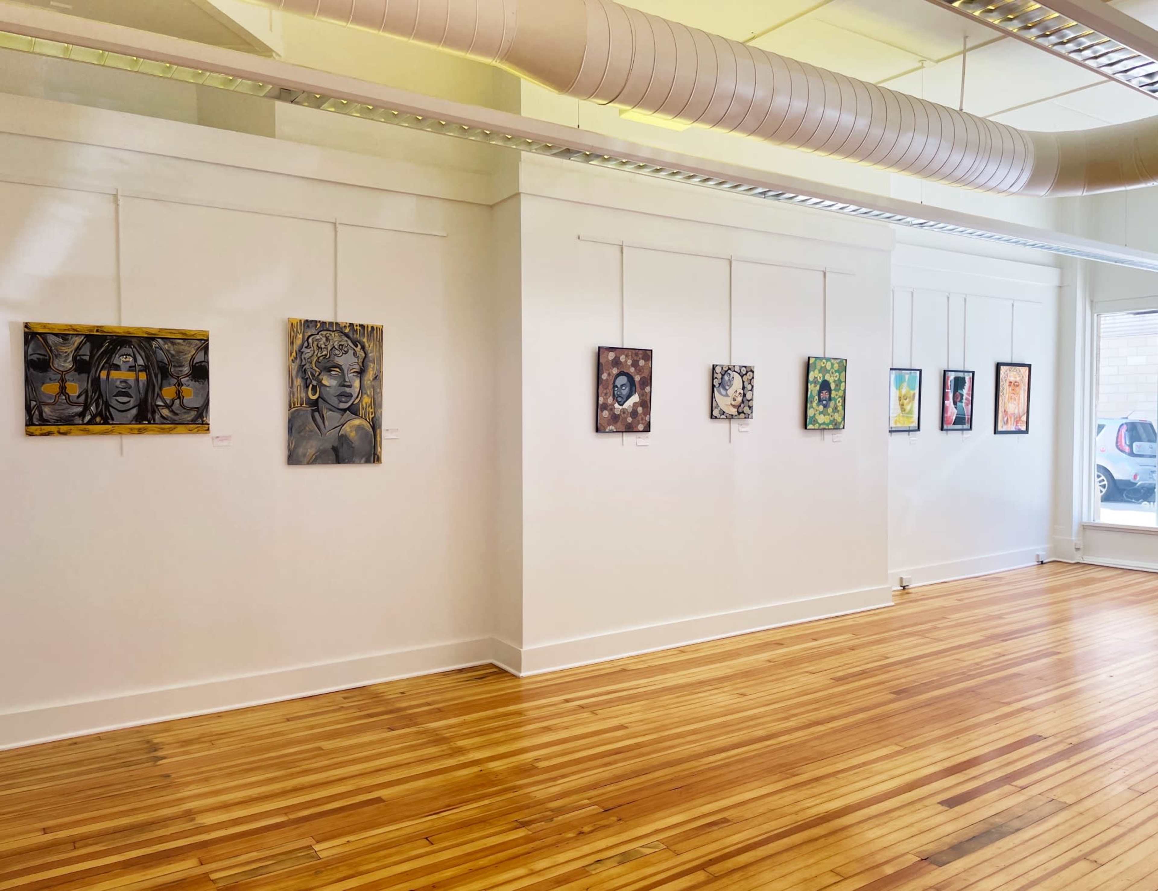 An art gallery wall displaying a series of framed artworks arranged in two rows on a white wall, with wooden flooring visible beneath.